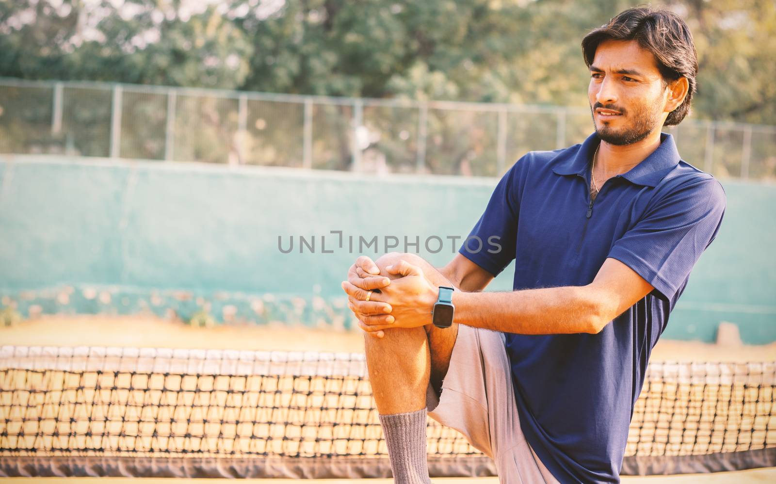 Young tennis player stretches legs before playing at tennis court - concept of warming up before playing any sports