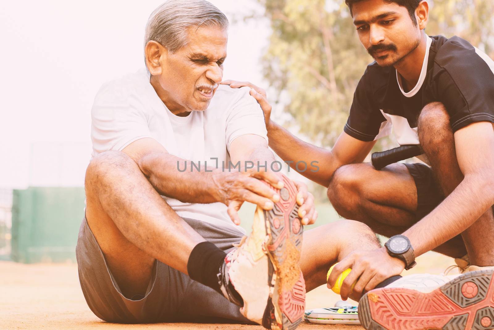 Elderly holding his toe suffering from pain and young man or teammate helping him at tennis court - concept of old people fitness and injury - young people helping senior citizens at sports. by lakshmiprasad.maski@gmai.com