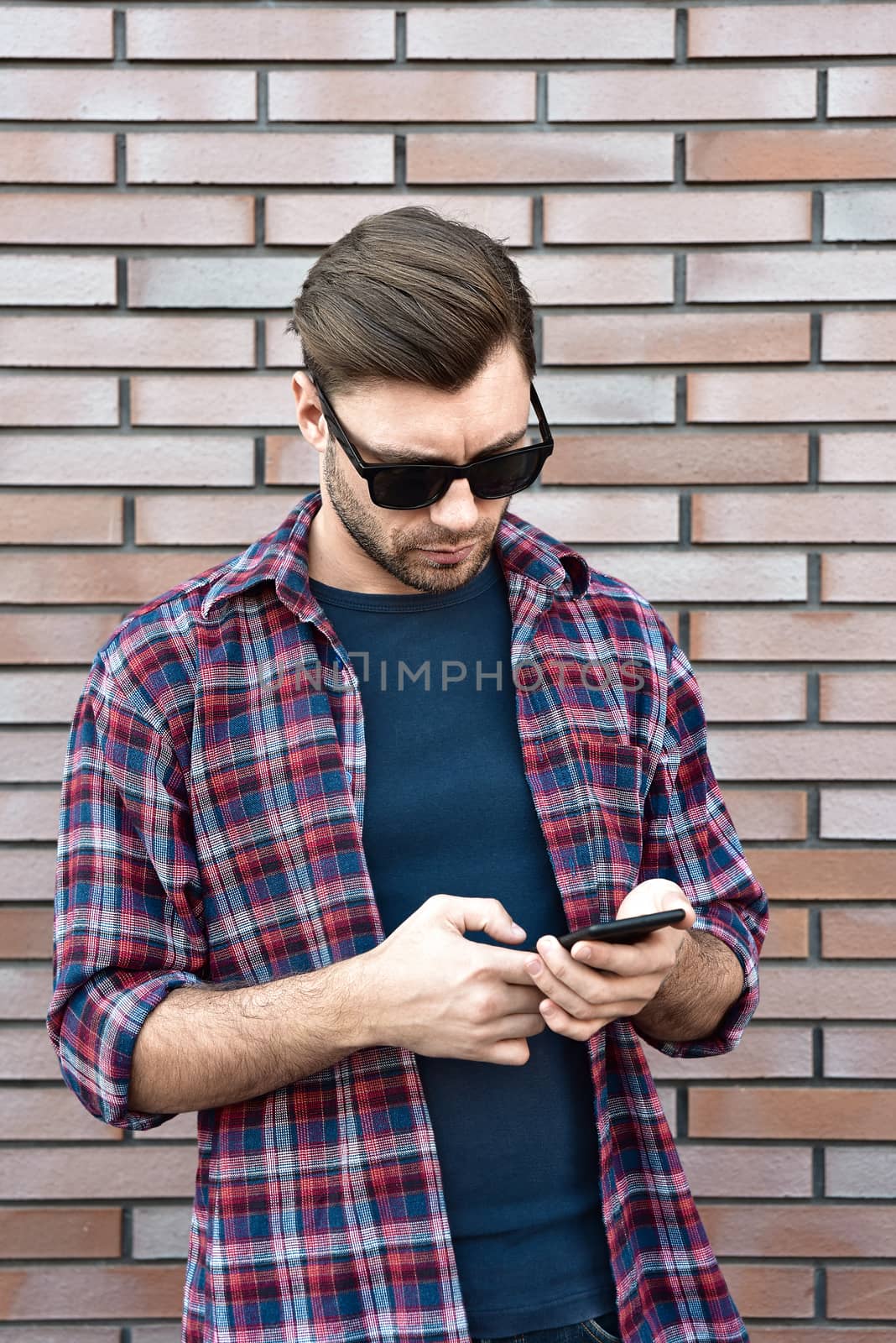 Front view of handsome young man in smart casual wear hold a mobile phone while standing at the brick wall background.