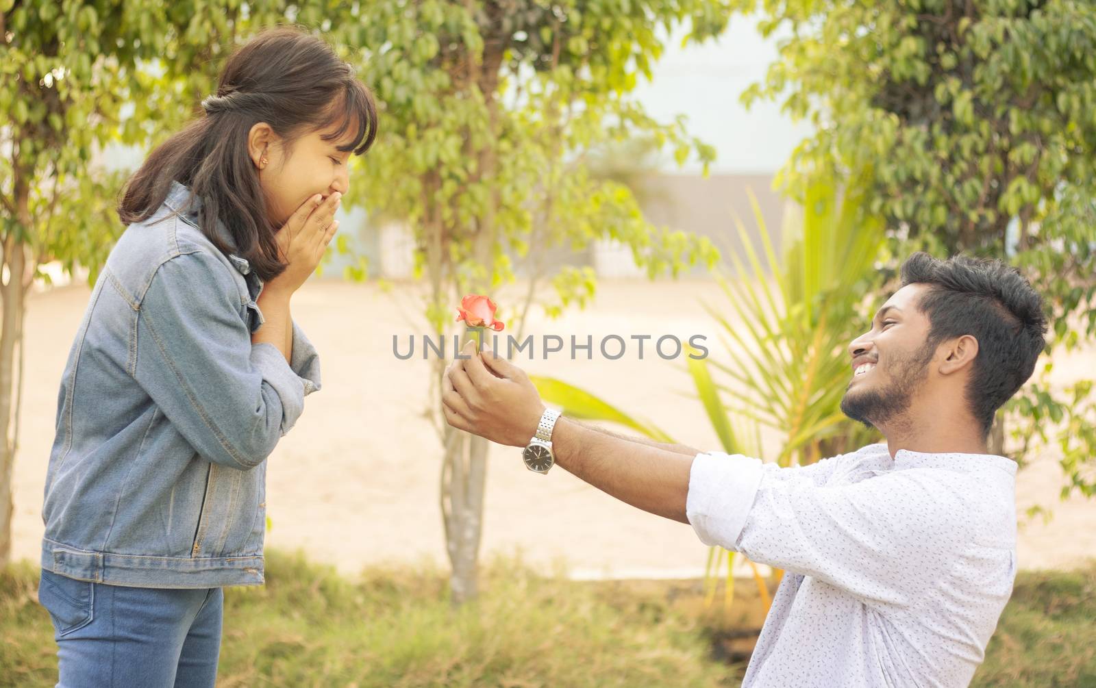 Concept of teenage love or affection - young man proposing to smiling excited girlfriend standing on knee with red rose on valentines day.