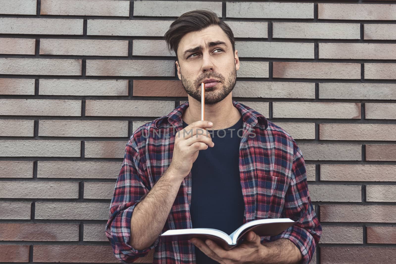 Young hipster guy wearing eyeglasses reading a book or note book make some notes and ideas on brick wall background.