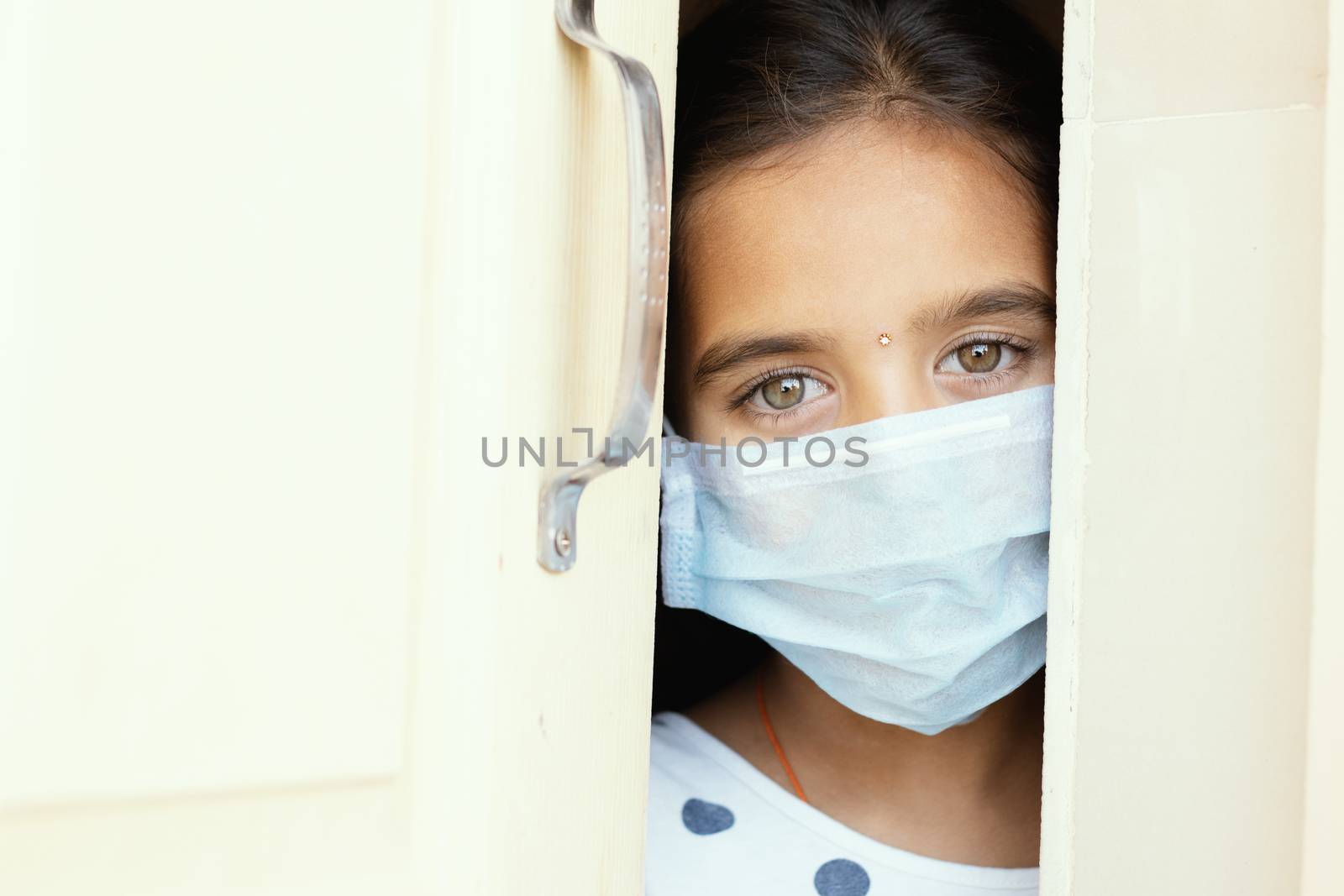 Young sad girl with medical mask wearing sneaking out through the home door - concept of home quarantine due to covid-19 or coronavirus outbreak