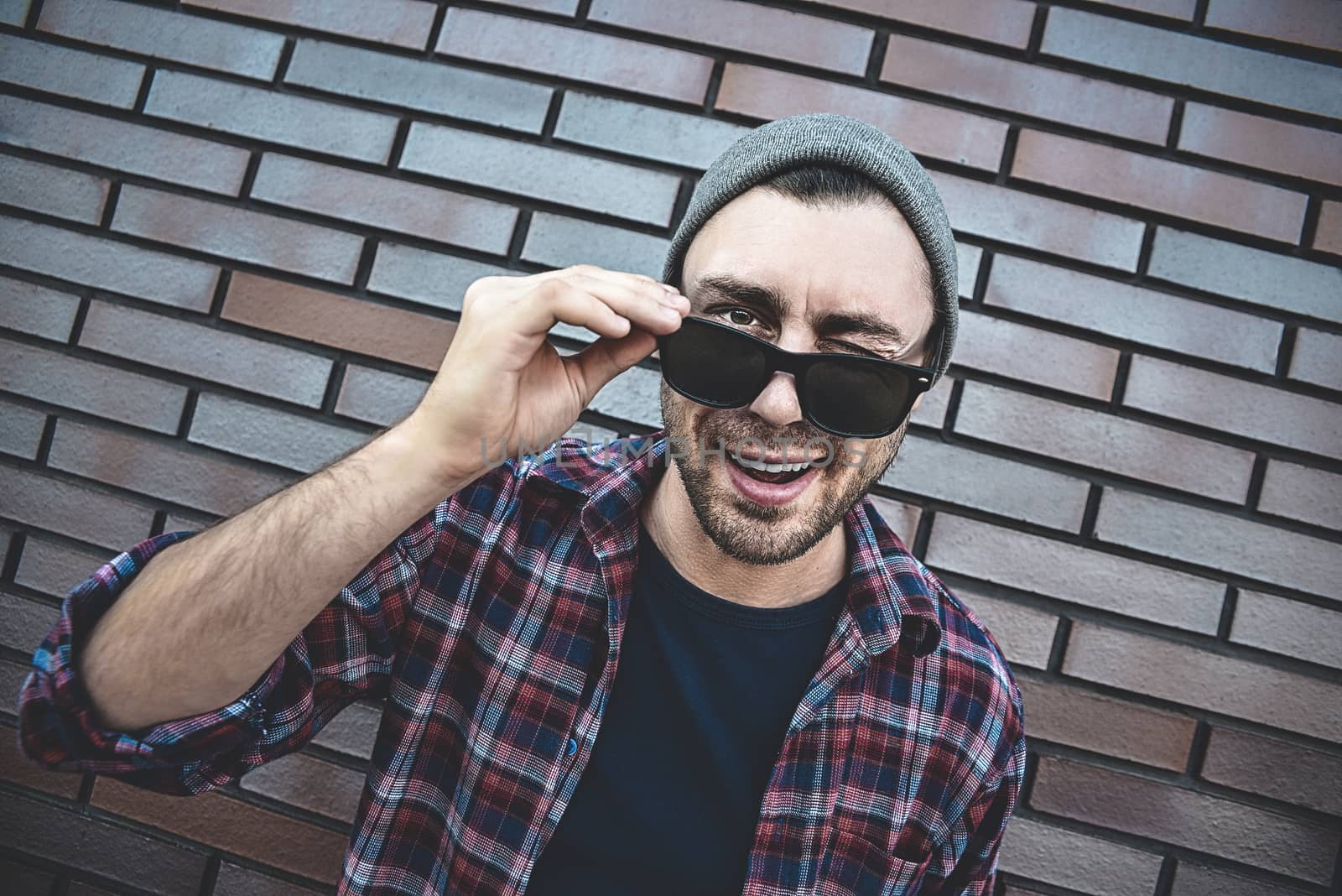 Young adult man wearing sunglasses standing over brown brick wall smiling with happy face winking at the camera.