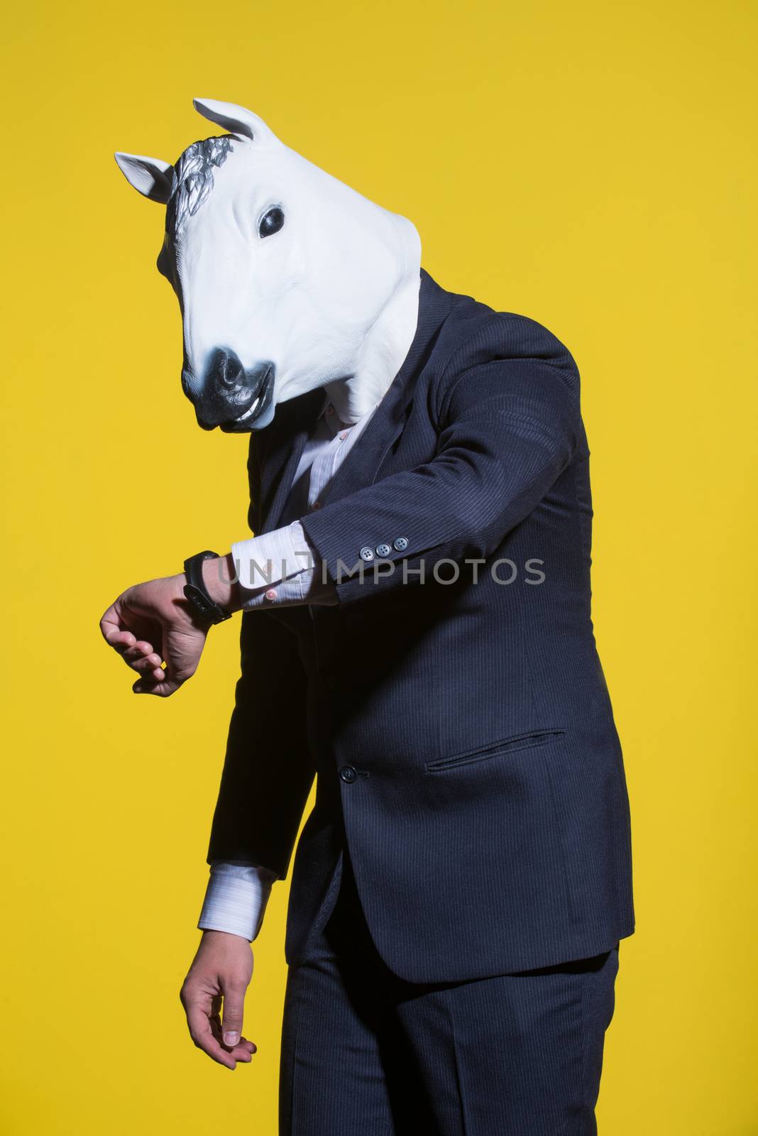 A man in a suit and a horse mask looking at his watch. Yellow background. Conceptual business background