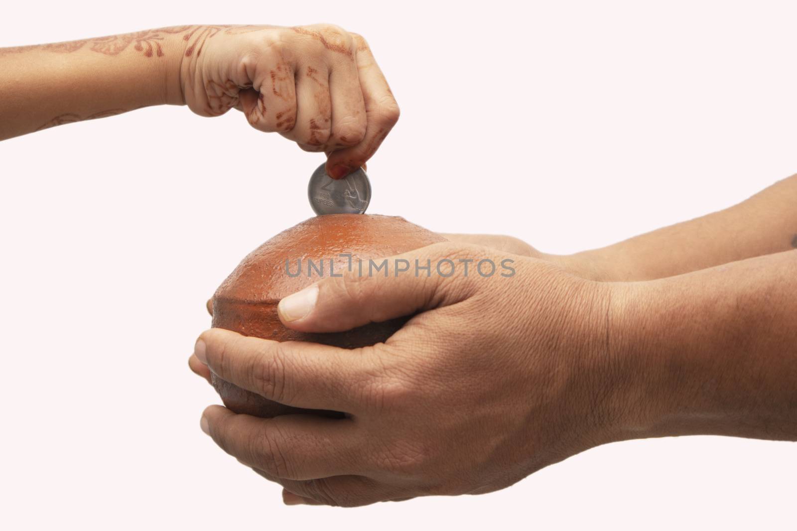 Hands Holding Indian penny bank made with mud clay and Child hand adding coin into the earthen pot or terracotta piggy bank on Isolated background - Concept showing of savings, investment or donating