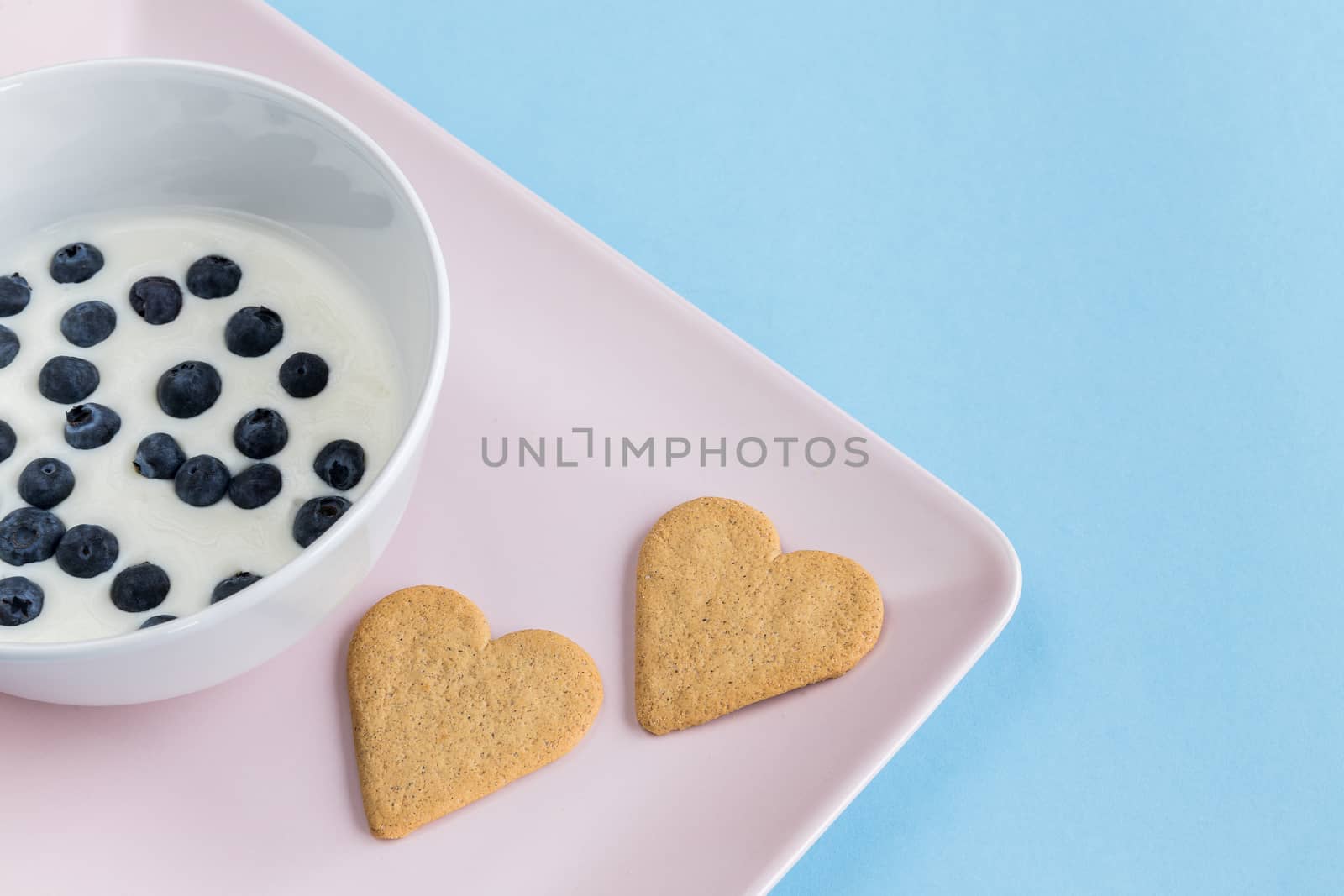 Heart-shaped cookies and a bowl of yogurt with blueberries on a pink tray on a light blue background. Copy space.