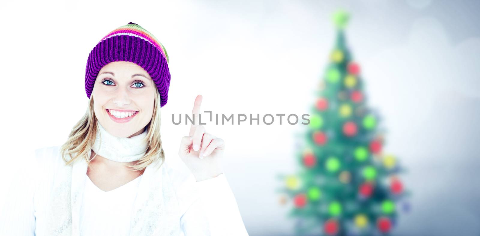 Joyful woman with a colorful hat pointing upwards against blurry christmas tree in room