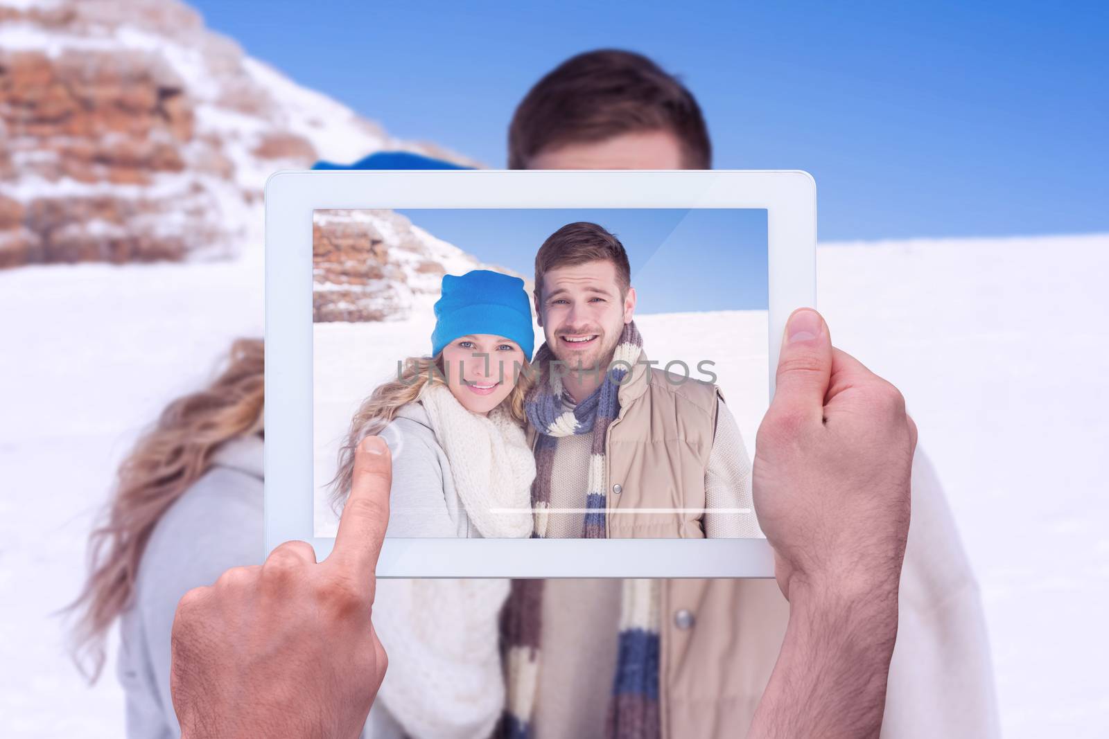 Hand holding tablet pc against couple in warm clothing on snow covered landscape