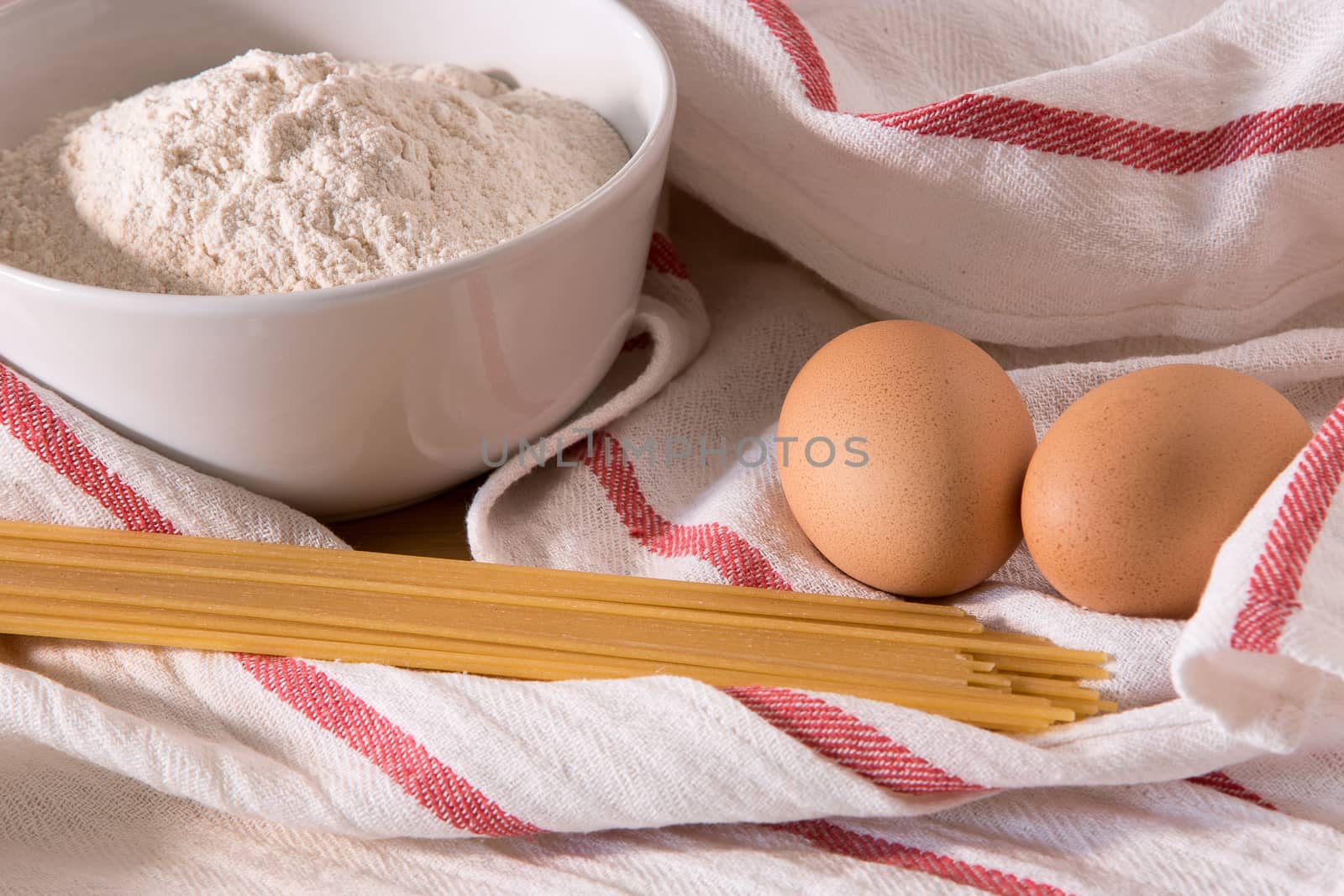 Gourmet close up with a bowl full of flour, spaghetti, and two eggs in white kitchen cloths with red border.