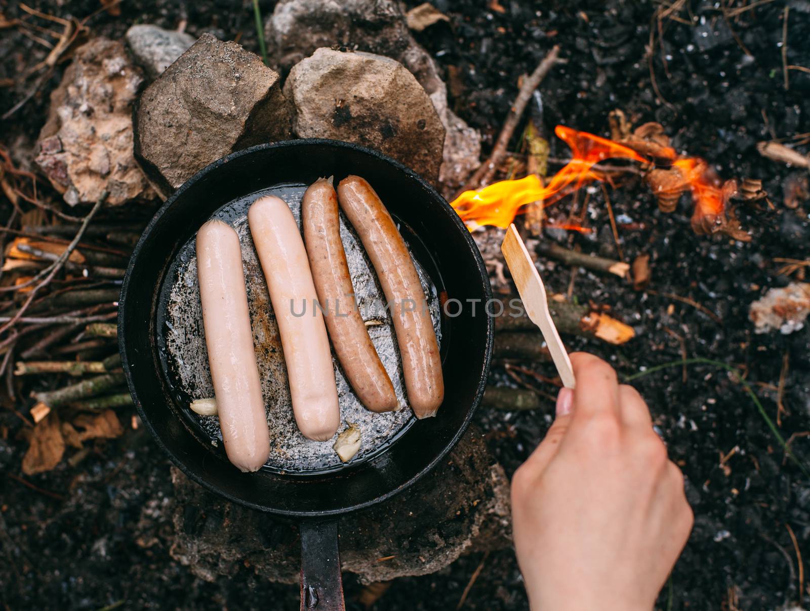 Roasting sausages in a frying pan over an open fire. Preparing food in nature. Lunch in the open air. Hand with a wooden shovel. Picnic in the forest. Food on fire