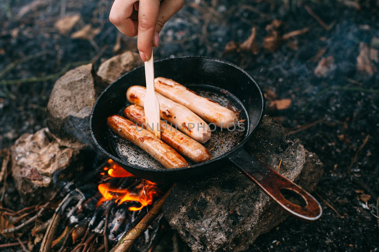 Roasting sausages in a frying pan over an open fire. Preparing food in nature. Lunch in the open air. Hand with a wooden shovel. Picnic in the forest. Food on fire