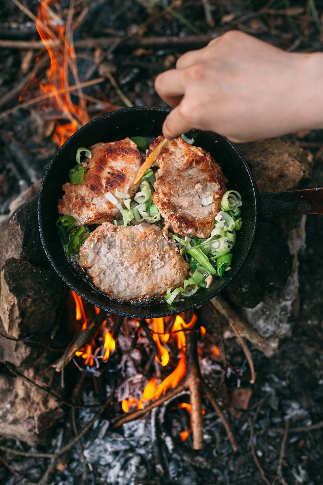 Frying meat in a pan over an open fire with leek. Steak in a pan on a fire. Cooking in nature. Picnic. Grill on fire. Hand with a wooden spatula.