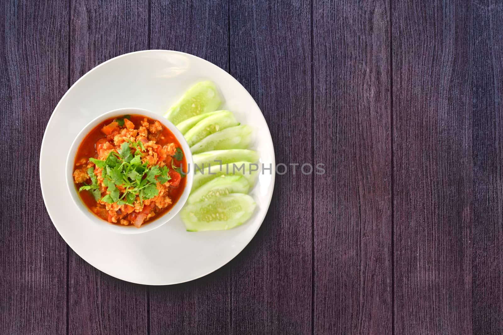 Thai Northern Style Pork and Tomato Relish in white bowl served with cucumber sliced on dark wooden background. Hot and spicy dipping. Top view.