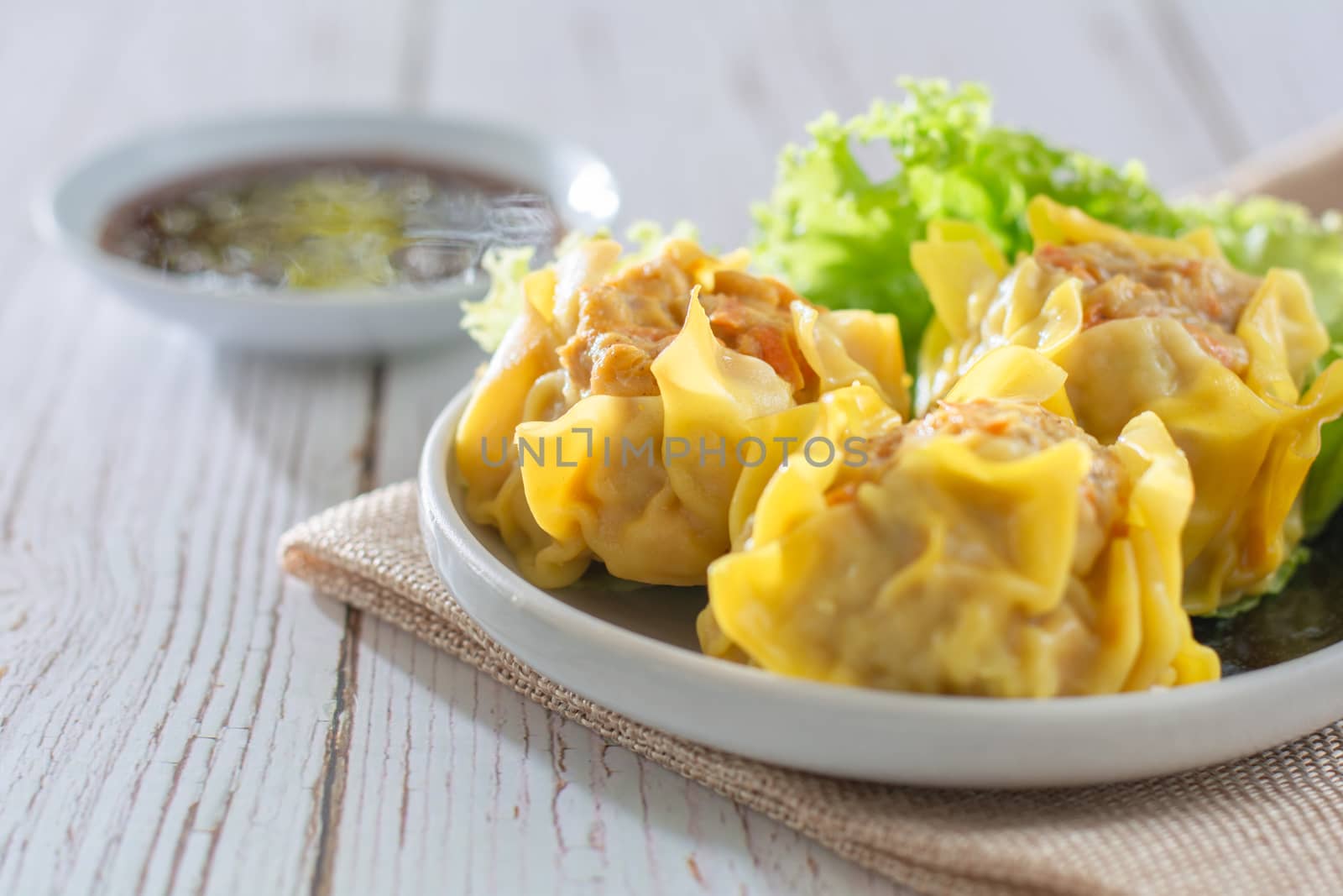 Chinese Steamed Dumpling, Shumai on white dish served with soy sauce and 
lettuce leaves on brown cloth and wooden table. Delicious Dimsum pork.