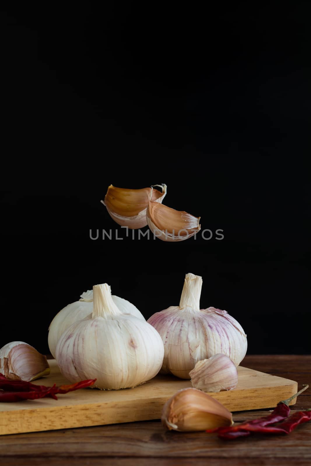 Group of garlic on chopping board and some garlic cloves floating in the air and red dried chilli on wooden table with black background. Copy space for your text.