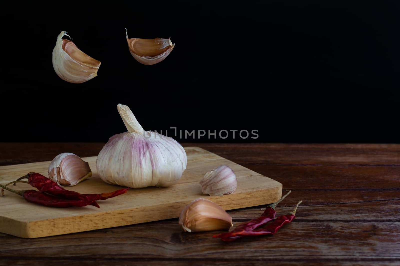 Group of garlic on chopping board and some garlic cloves floating in the air and red dried chilli on wooden table with black background. Copy space for your text.