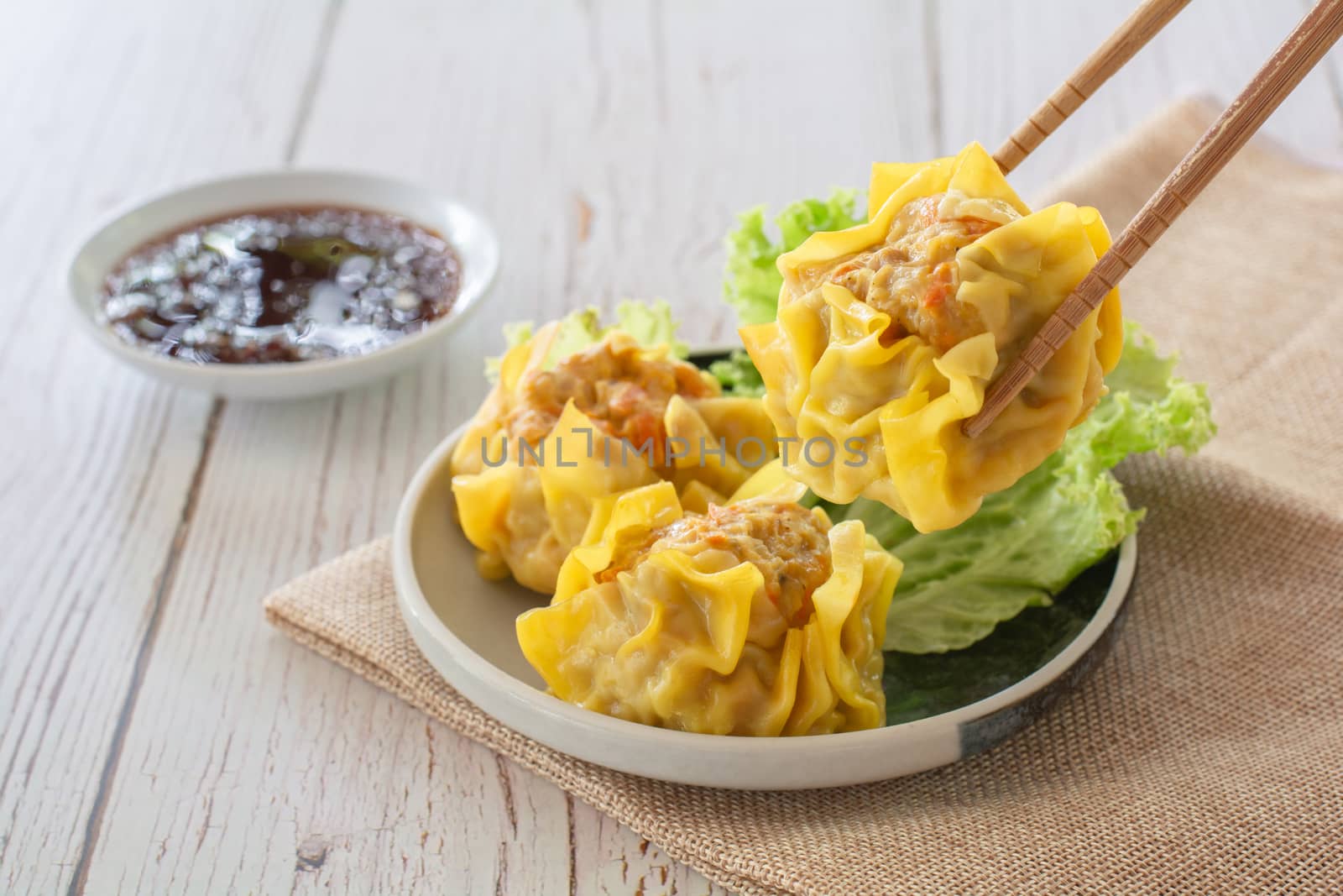 Chinese Steamed Dumpling, Shumai on white dish served with soy sauce and 
lettuce leaves on brown cloth and wooden table. Delicious Dimsum pork.