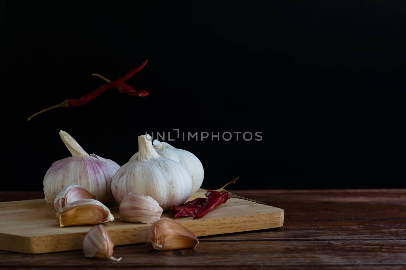 Group of garlic on chopping board and some garlic cloves floating in the air and red dried chilli on wooden table with black background. Copy space for your text.