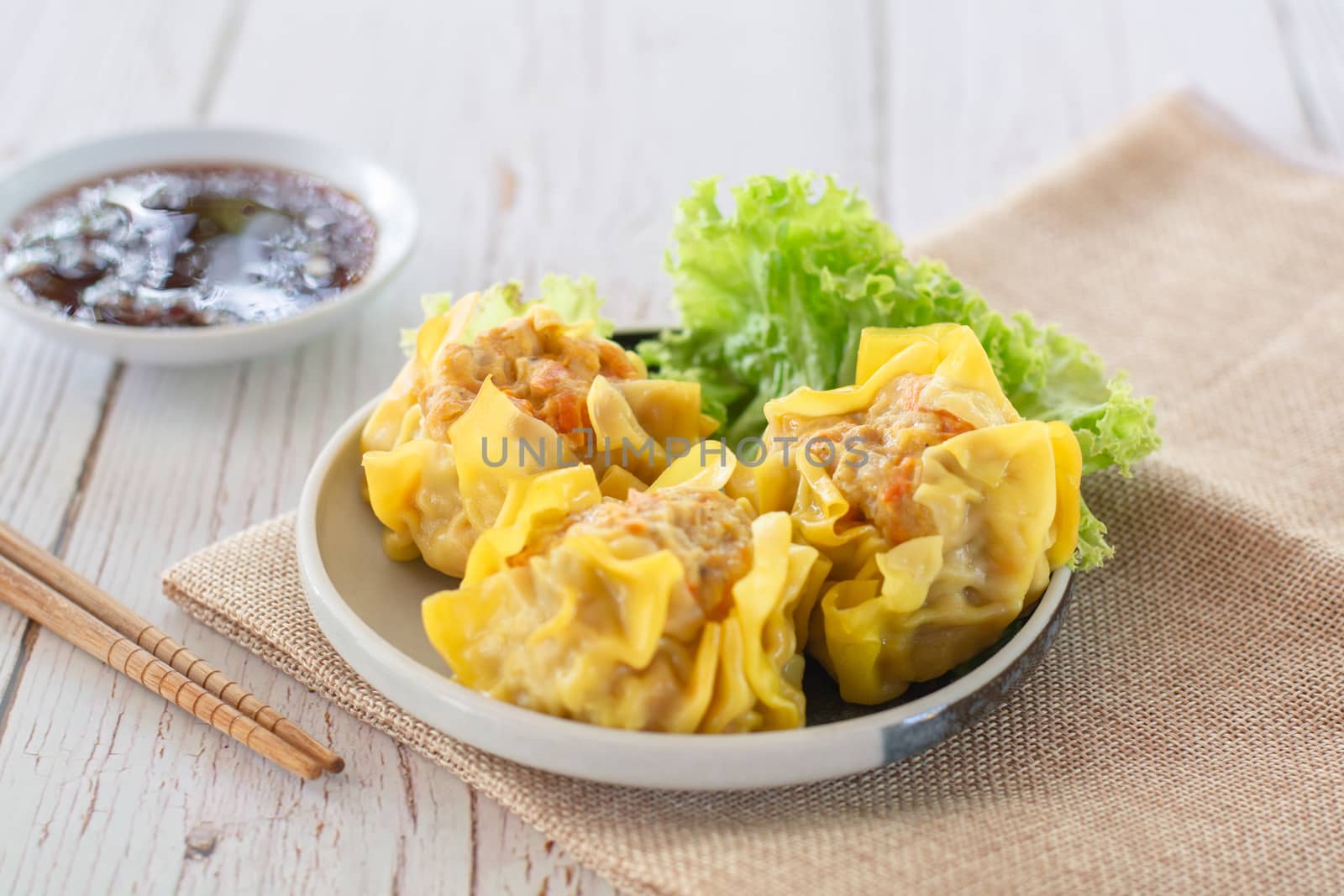 Chinese Steamed Dumpling, Shumai on white dish served with soy sauce and 
lettuce leaves on brown cloth and wooden table. Delicious Dimsum pork.