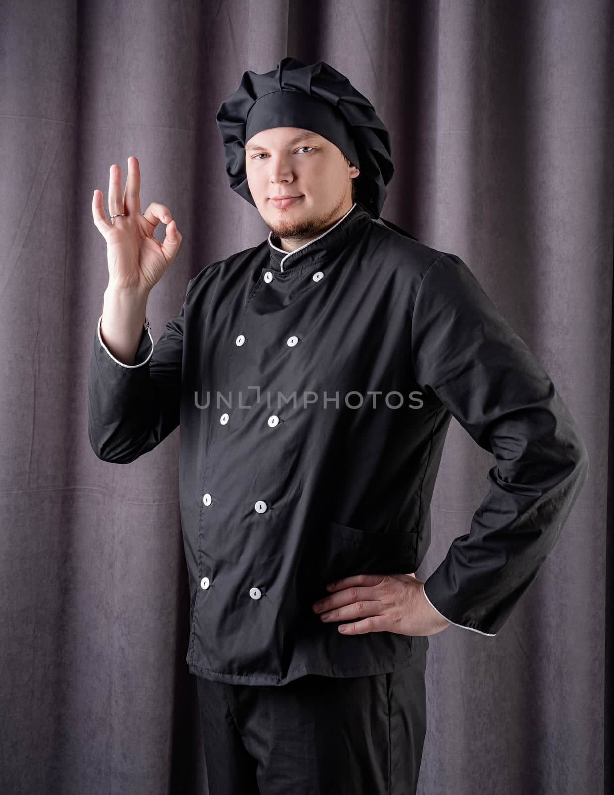 Young smiling male chef showing ok sign isolated on dark curtain background