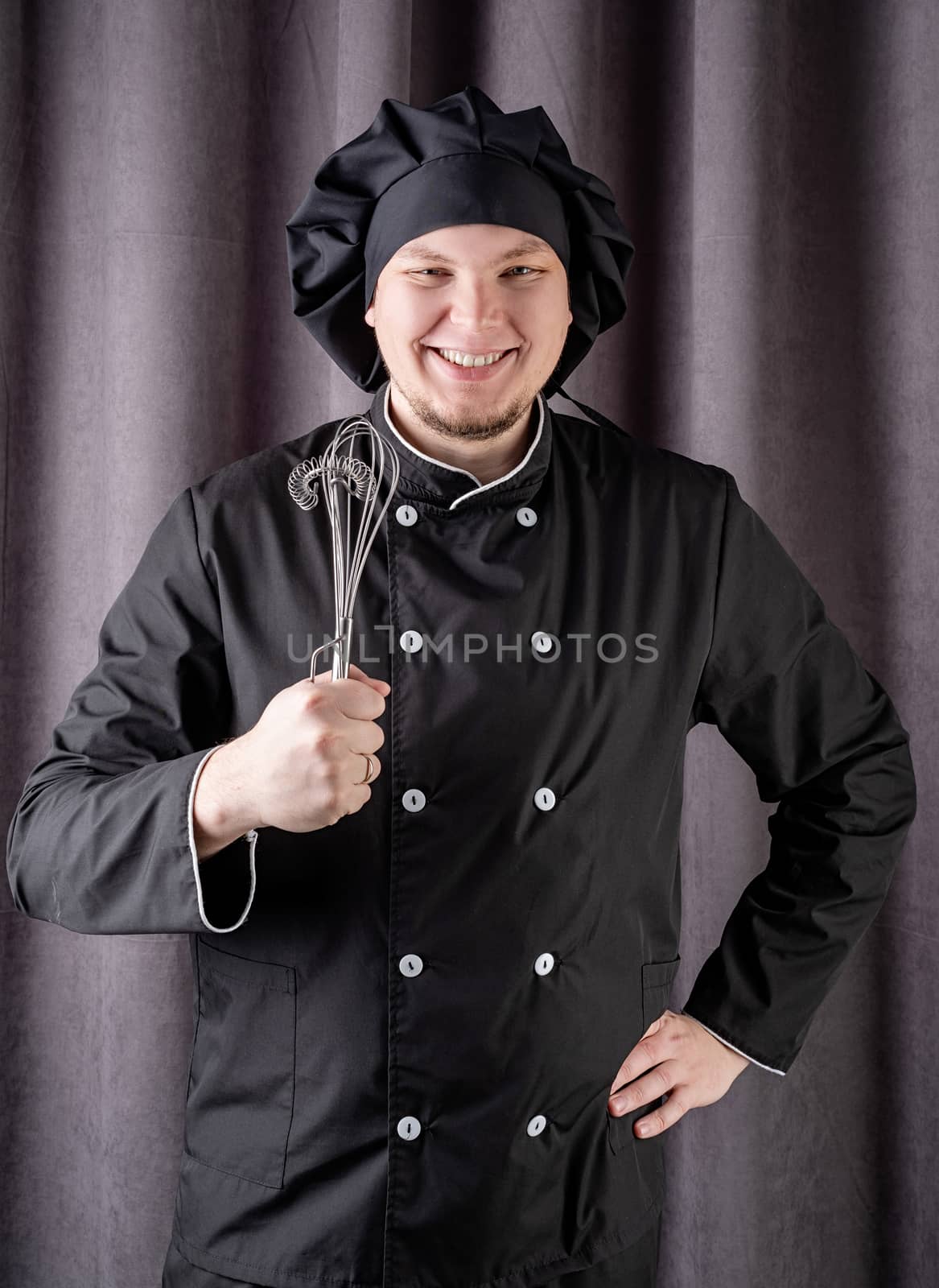 Young smiling male chef holding kitchen utencils isolated on dark curtain background
