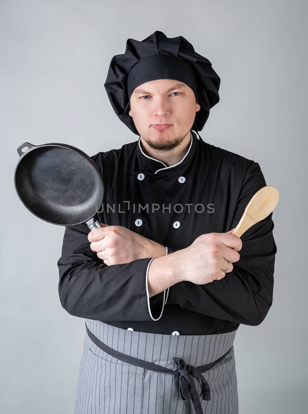 young chef in black uniform holding a saucepan and a spoon isolated on gray background by Desperada