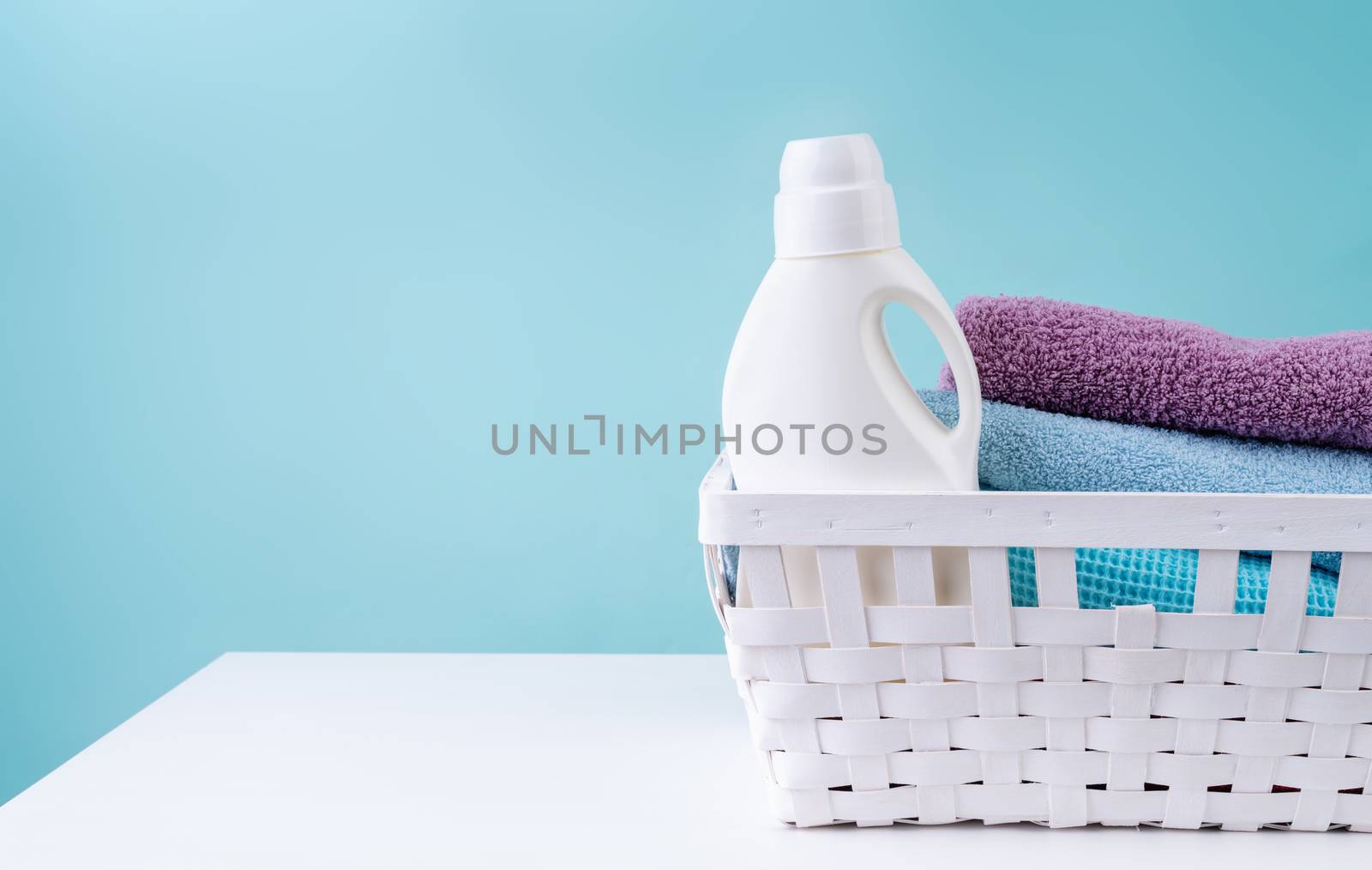 Laundry basket with a detergent bottle and a pile of clean towels on white table isolated on blue background by Desperada