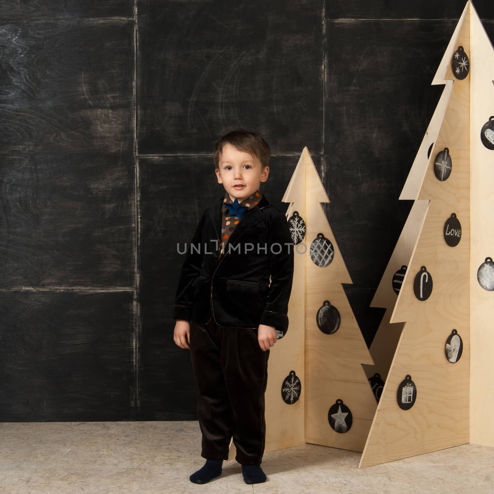 on a dark background little boy posing in a stylish costume near a Christmas tree made of wood