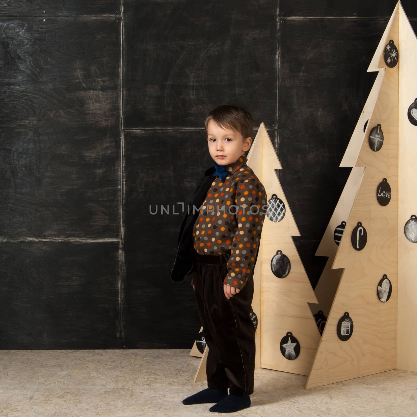 on a dark background little boy posing in a stylish costume near a Christmas tree made of wood