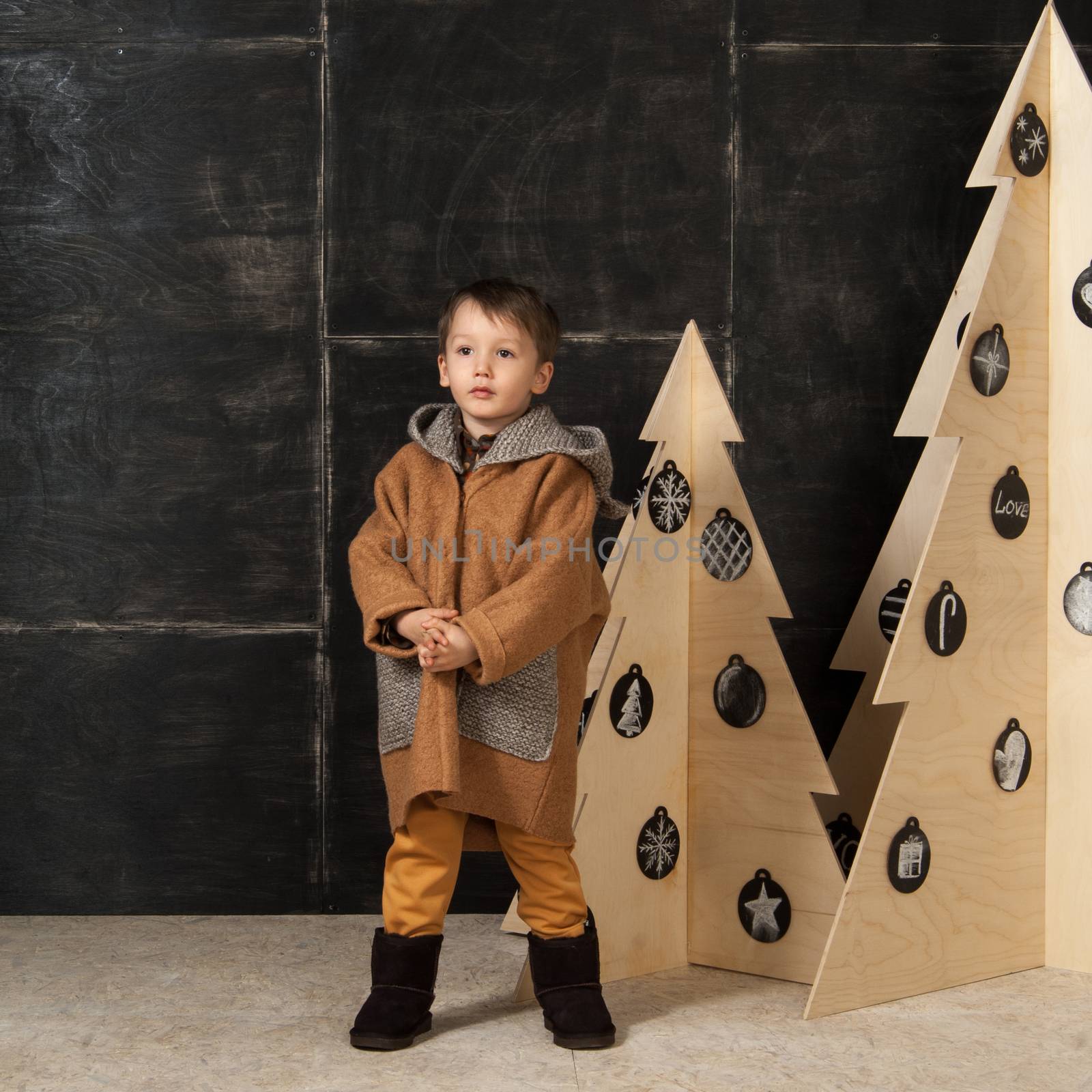 on a dark background little boy posing in a stylish coat near a Christmas tree made of wood