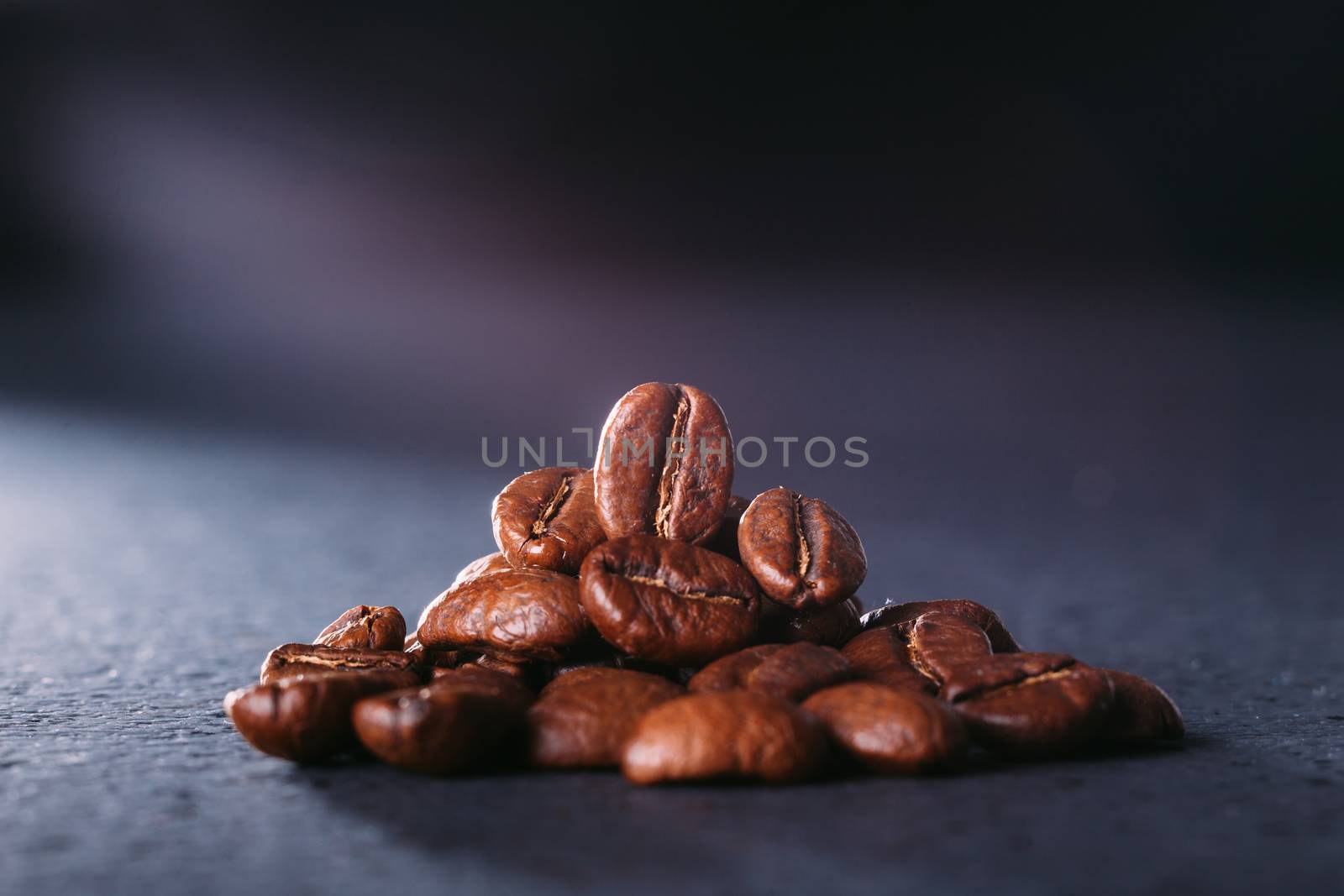 Grains of black coffee on a black granite countertop with yellow light from behind. Advertising photo of coffee. Photo with a glare from the light. Back light on a pyramid with grains.