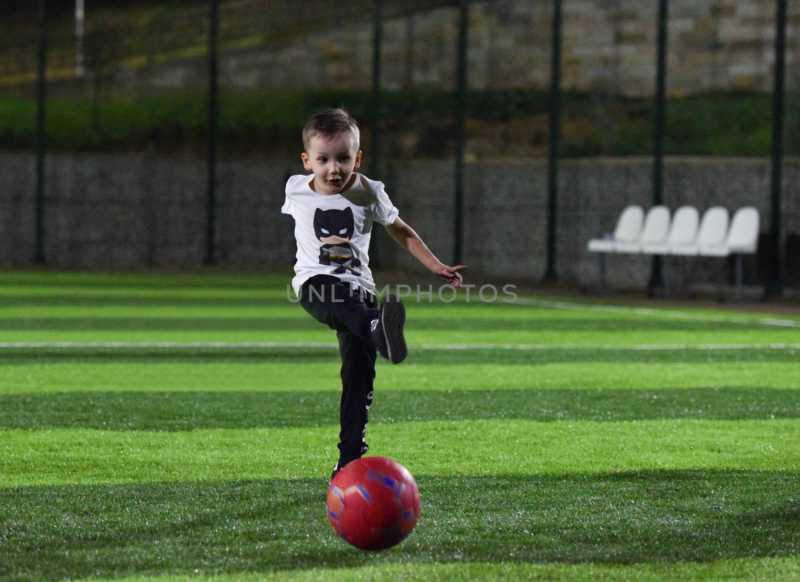 happy child kicks a red ball on the football field, night time