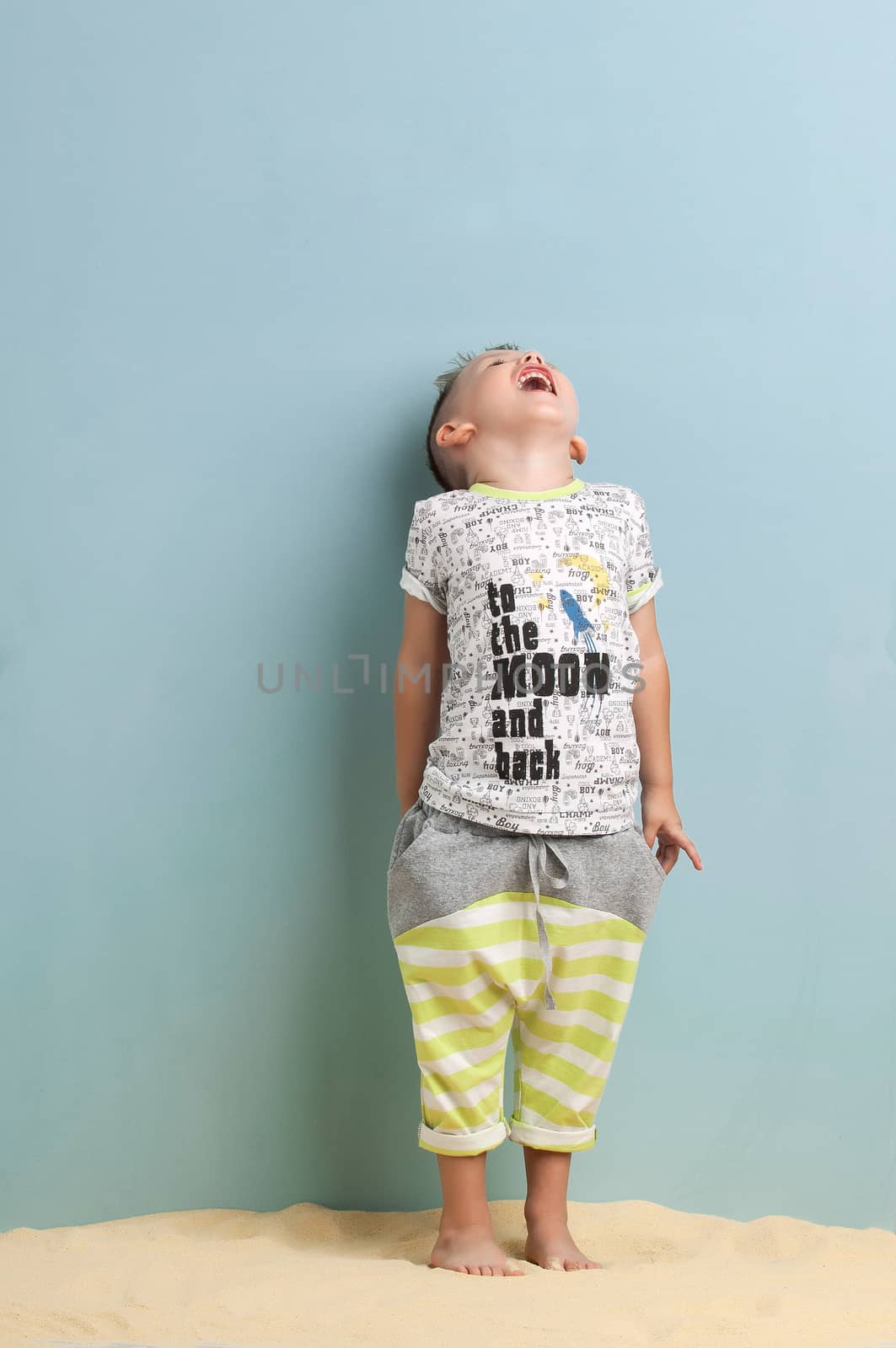 little boy in a shirt and shorts standing on the sand on a light blue background