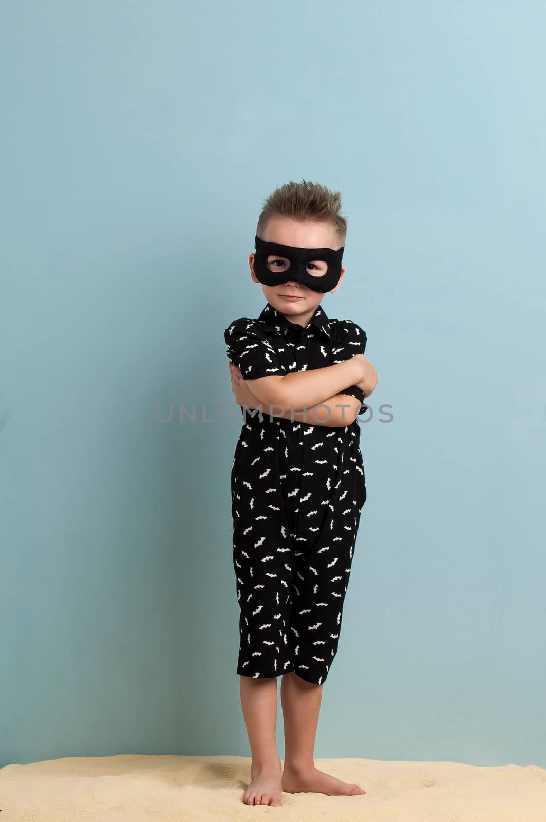 little boy in a black suit and a mask standing on sand on a light blue background