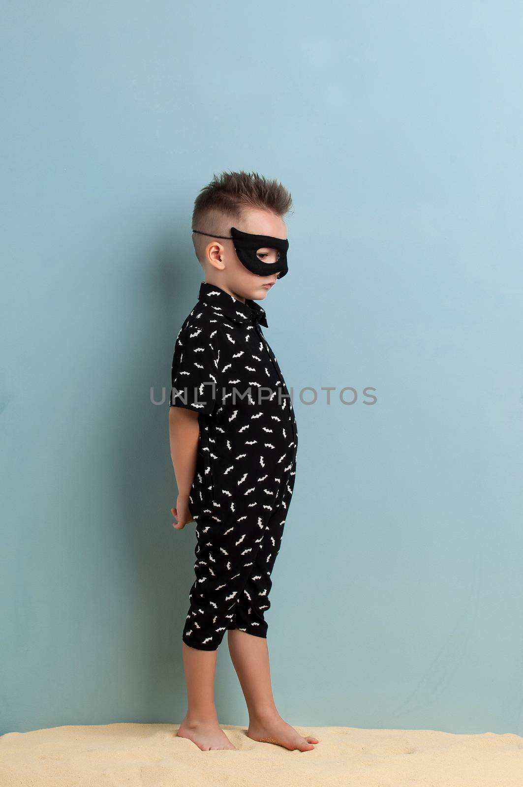 little boy in a black suit and a mask standing on sand on a light blue background
