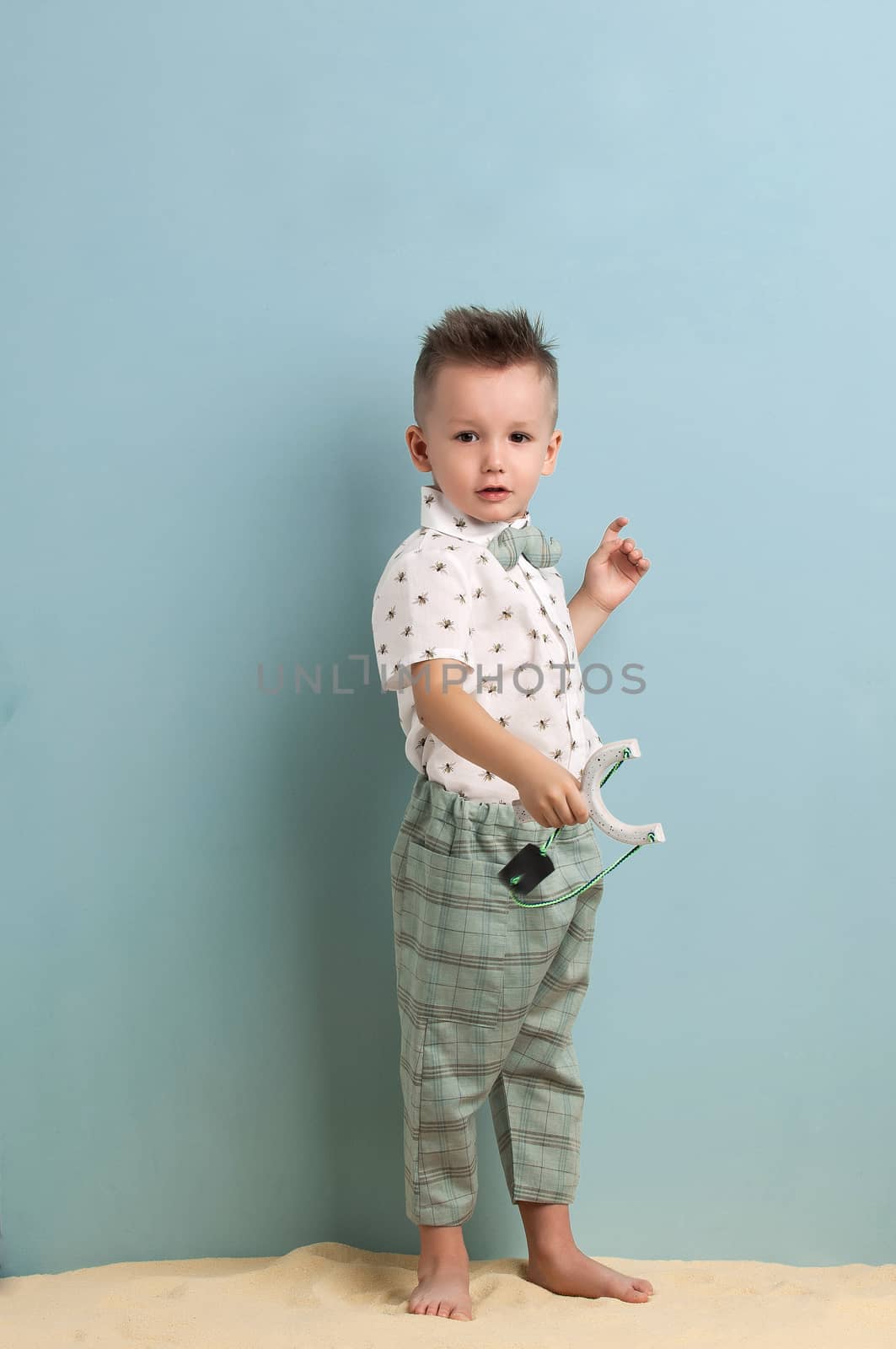 little boy in fashionable clothing and a slingshot in his hands stands on the sand on a light blue background