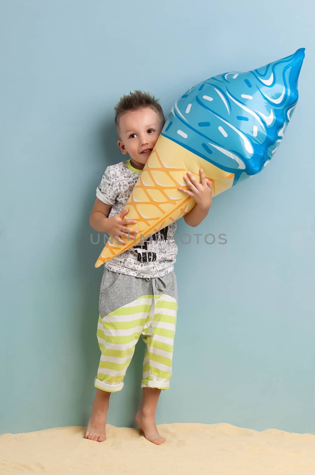little boy with an inflatable ice cream in the hands stands on the sand on a light blue background