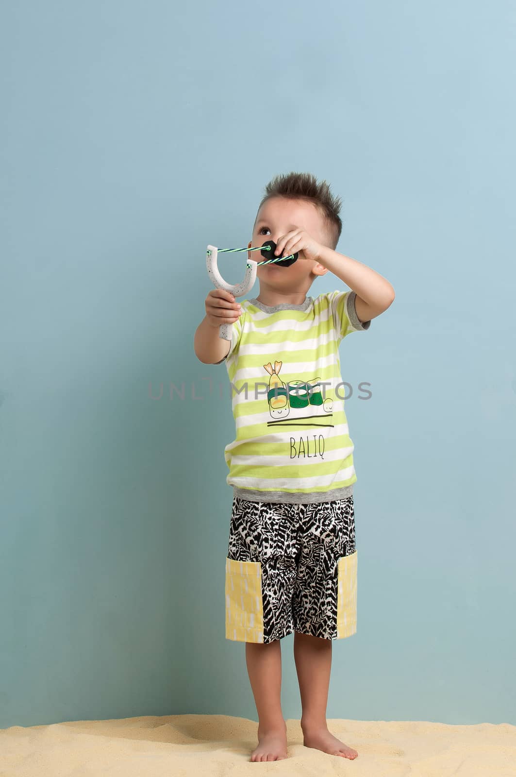 little boy in shorts and a T-shirt with a slingshot in his hands stands on the sand on a light blue background BALIQ - A FISH