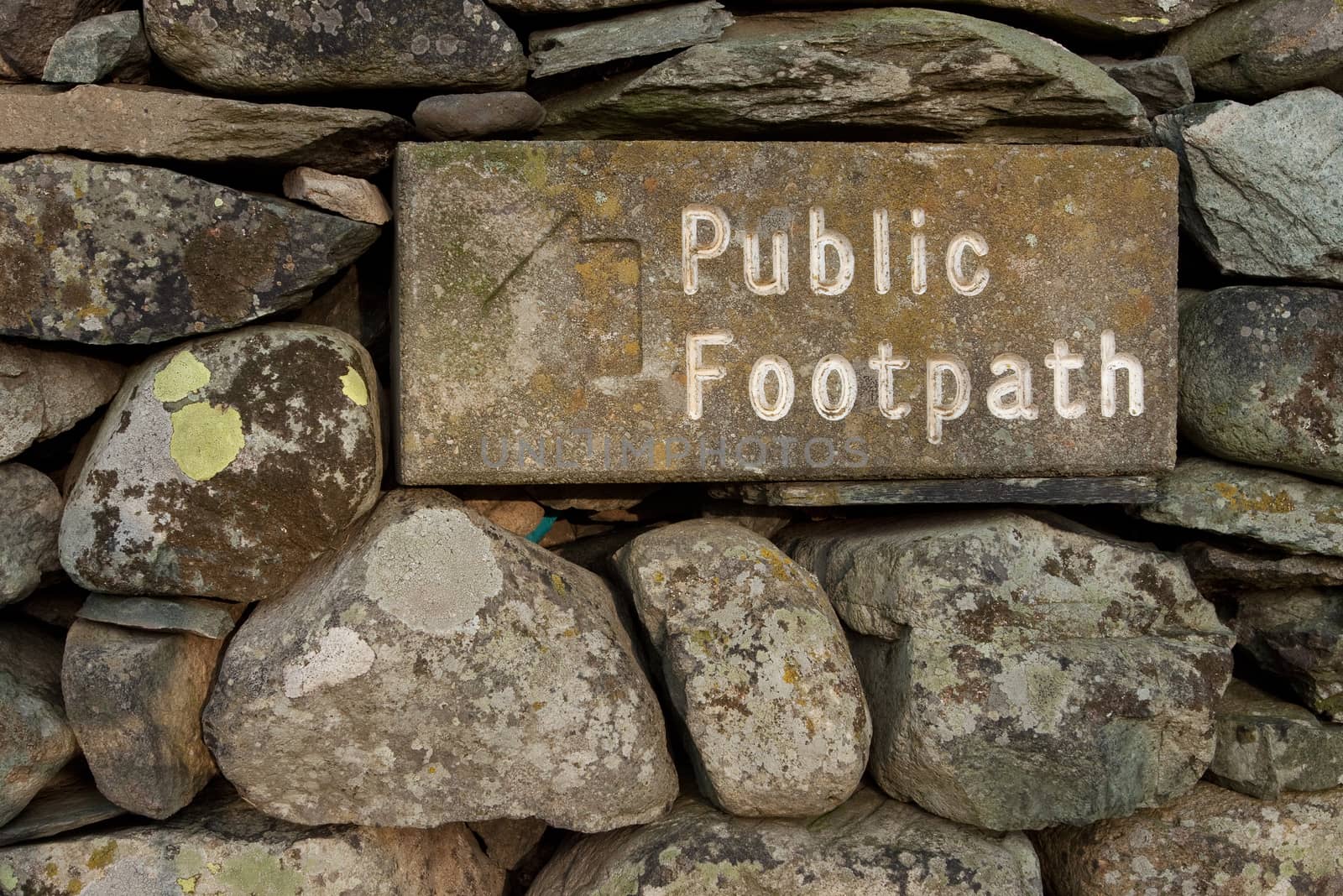 The stone public footpath sign is embedded in a dry stone wall and is situated alongside Ullswater lake in the English Lake District national park.