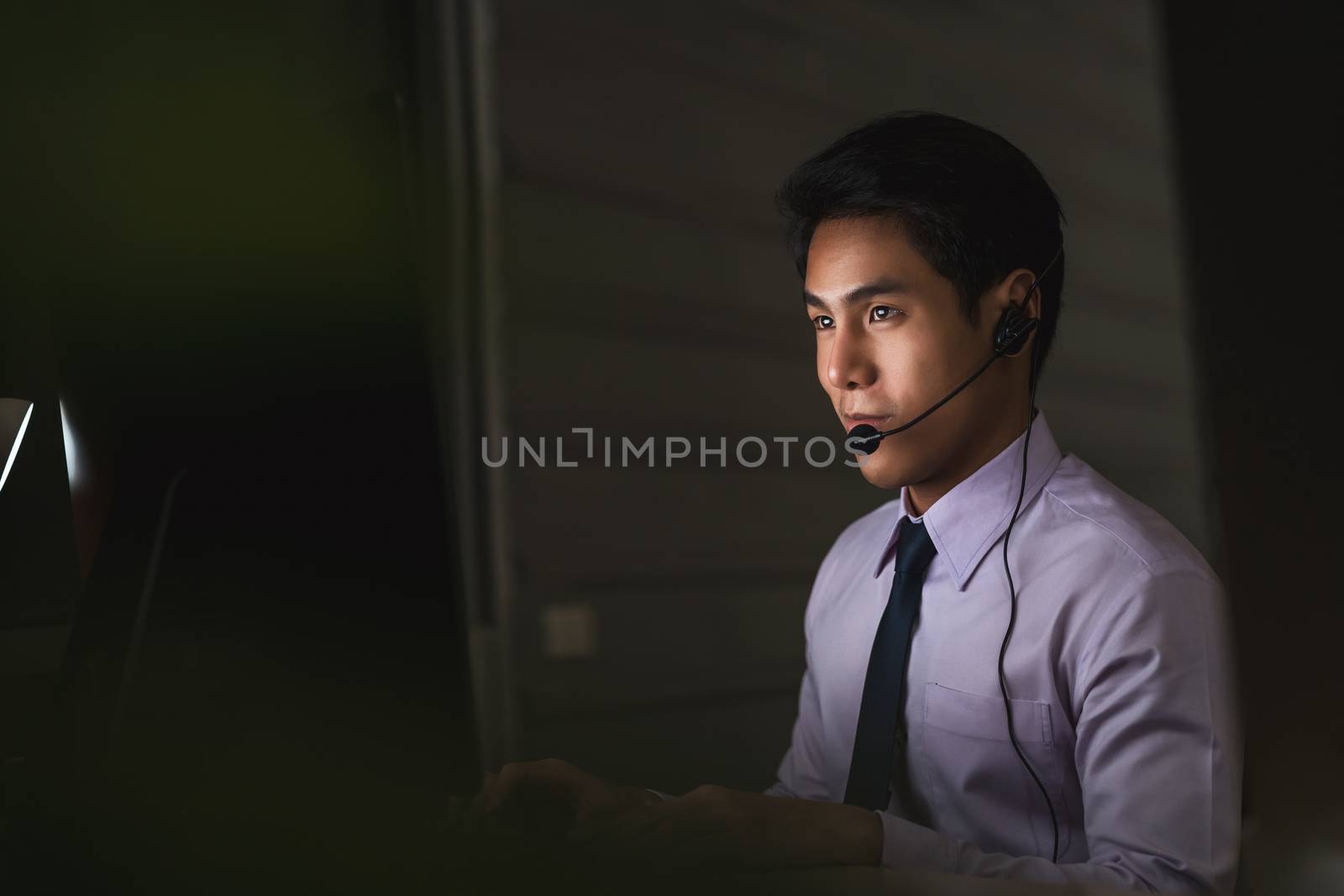 Asian male customer care service smiling and working hard late in night shift at office, call center department,worker and overtime, 7 days 24 hour support,teamwork with colleagues for success concept