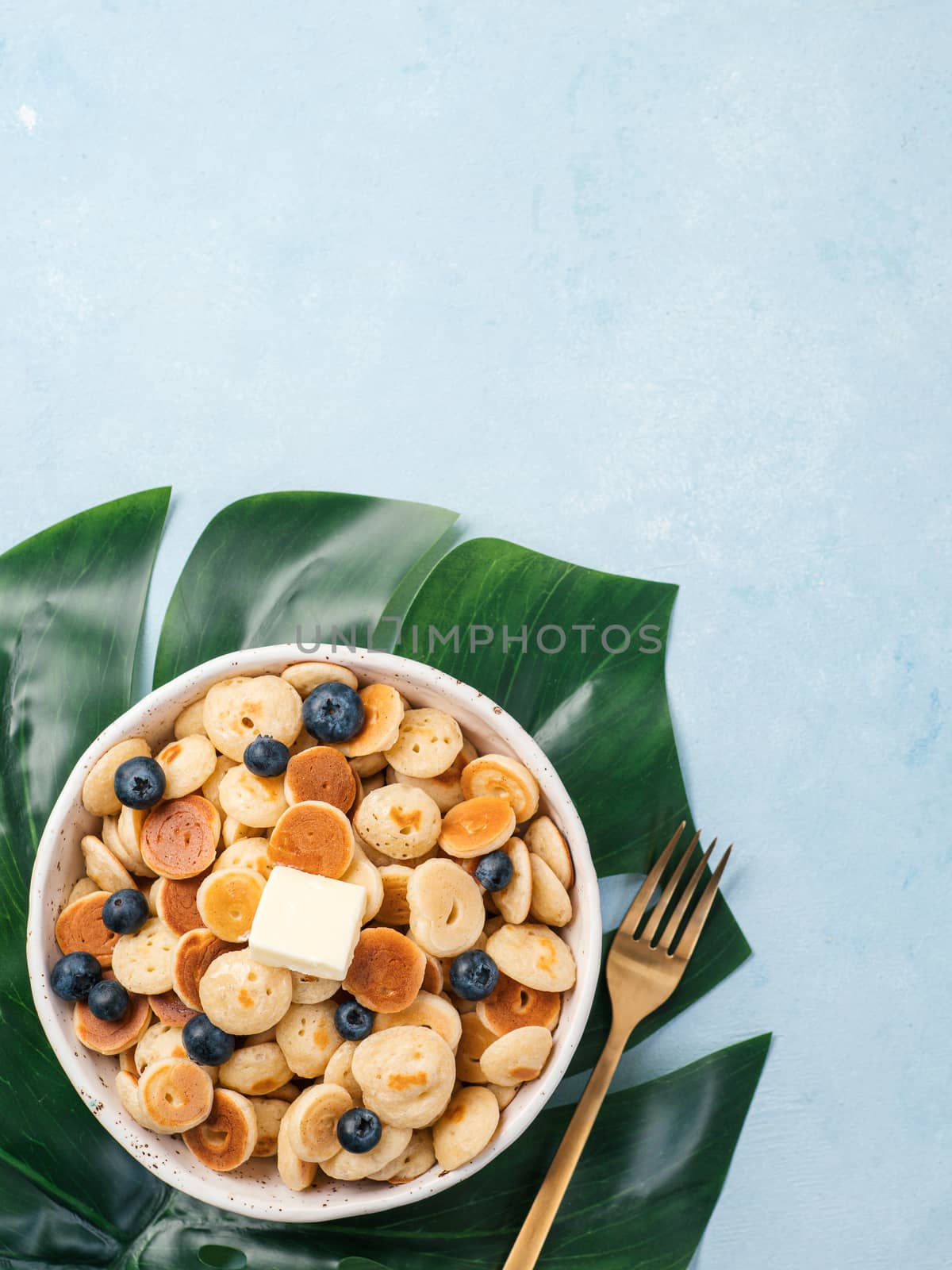 Trendy food - pancake cereal. Heap of mini cereal pancakes in boul on blue background. Copy space left for text. Top view or flat lay. Vertical.