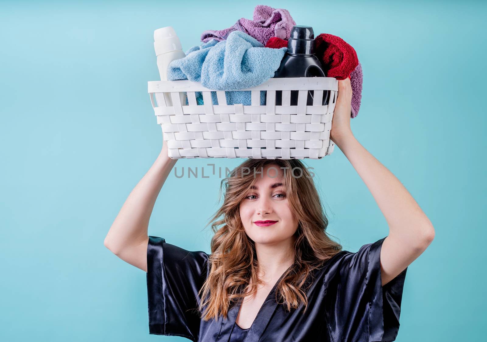Laundry concept. Happy housewife holding a basket of clothes ready for laundry on her head isolated on blue background with copy space