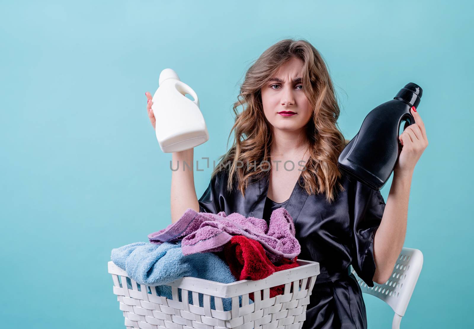 Laundry concept. puzzled housewife holding a basket of clothes and detergent bottles ready for laundry isolated on blue background with copy space