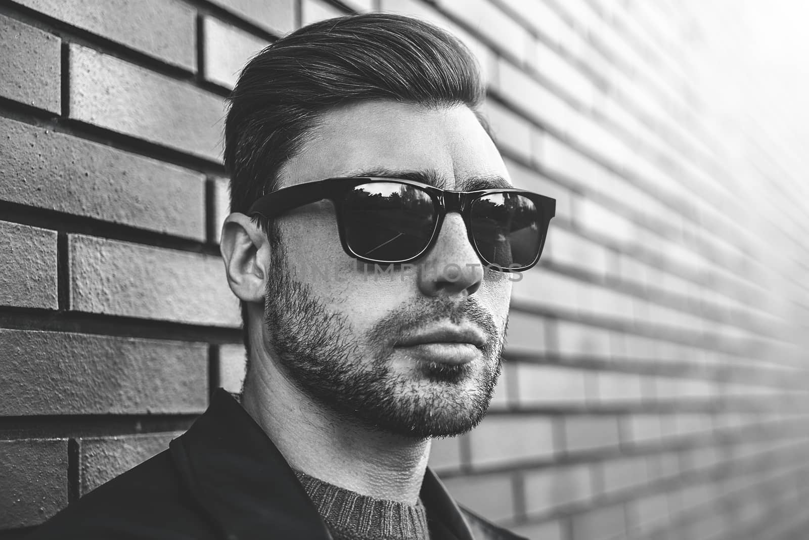 Portrait of stylish handsome young man with bristle standing outdoors and leaning on brick wall. Man wearing jacket, sweater and sunglasses.