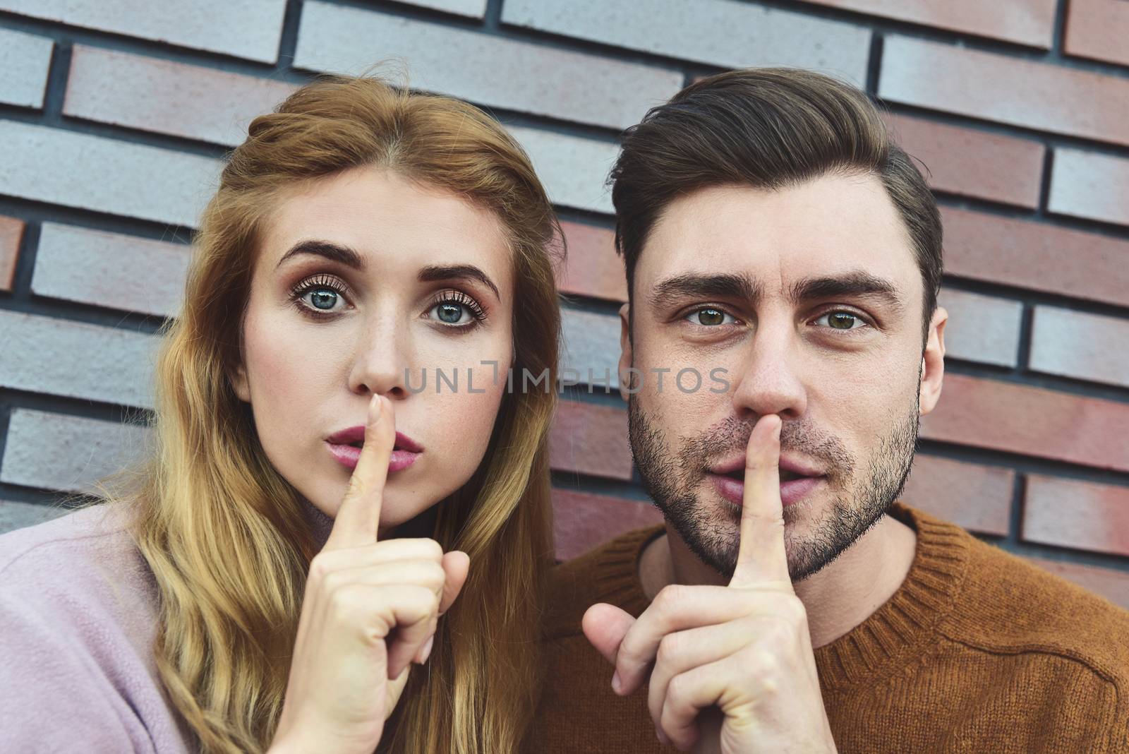 Shh, dont tell to anybody this private information. Surprised caucasian couple make shush gesture with surprised expressions, ask not spread gossips about colleagues, isolated over brick background. by Nickstock