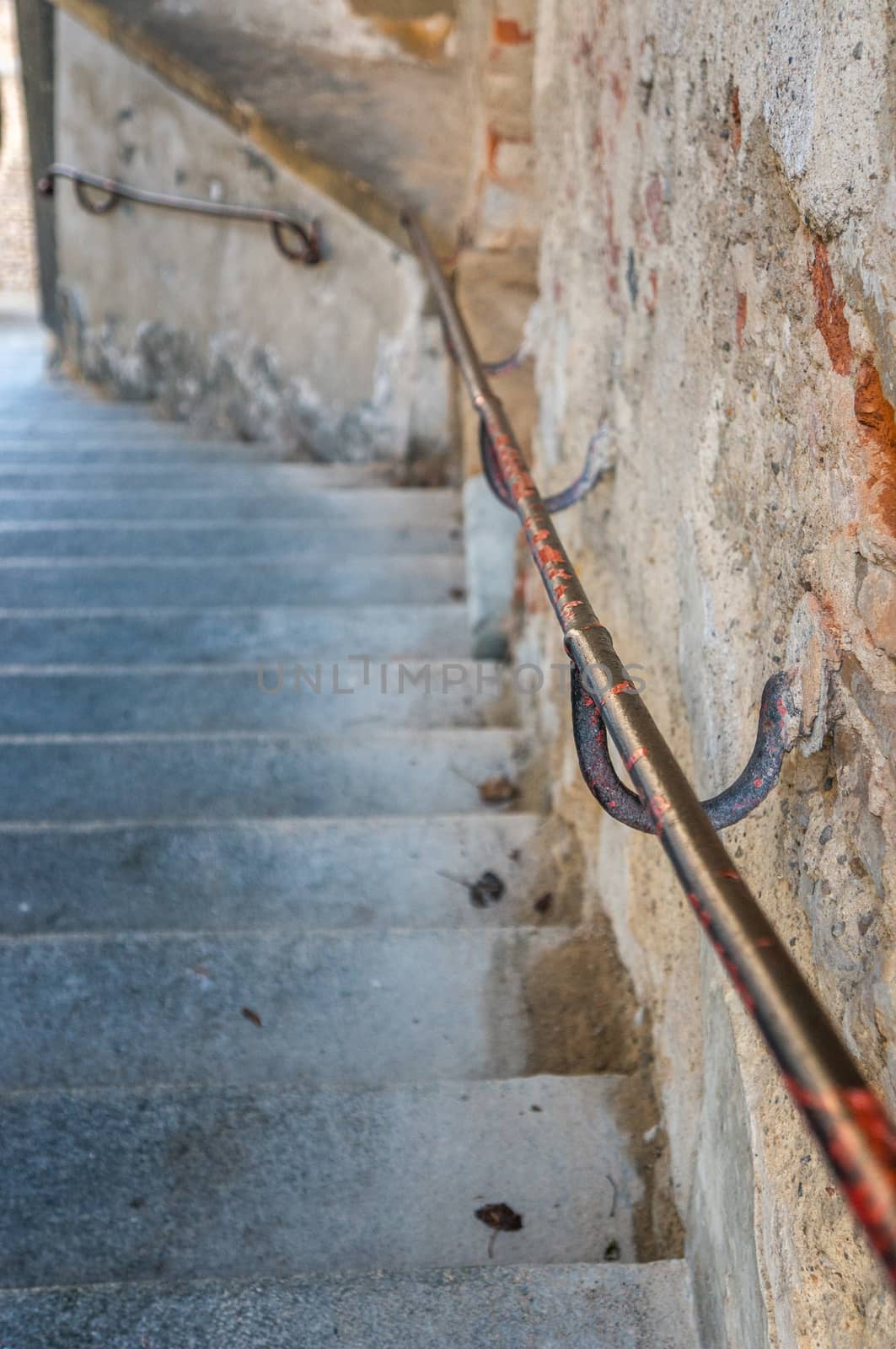 Close up shot of rust hand rail down stairs with red chipped off paint against worn stone wall. Stone steps in bright passage down a hill.