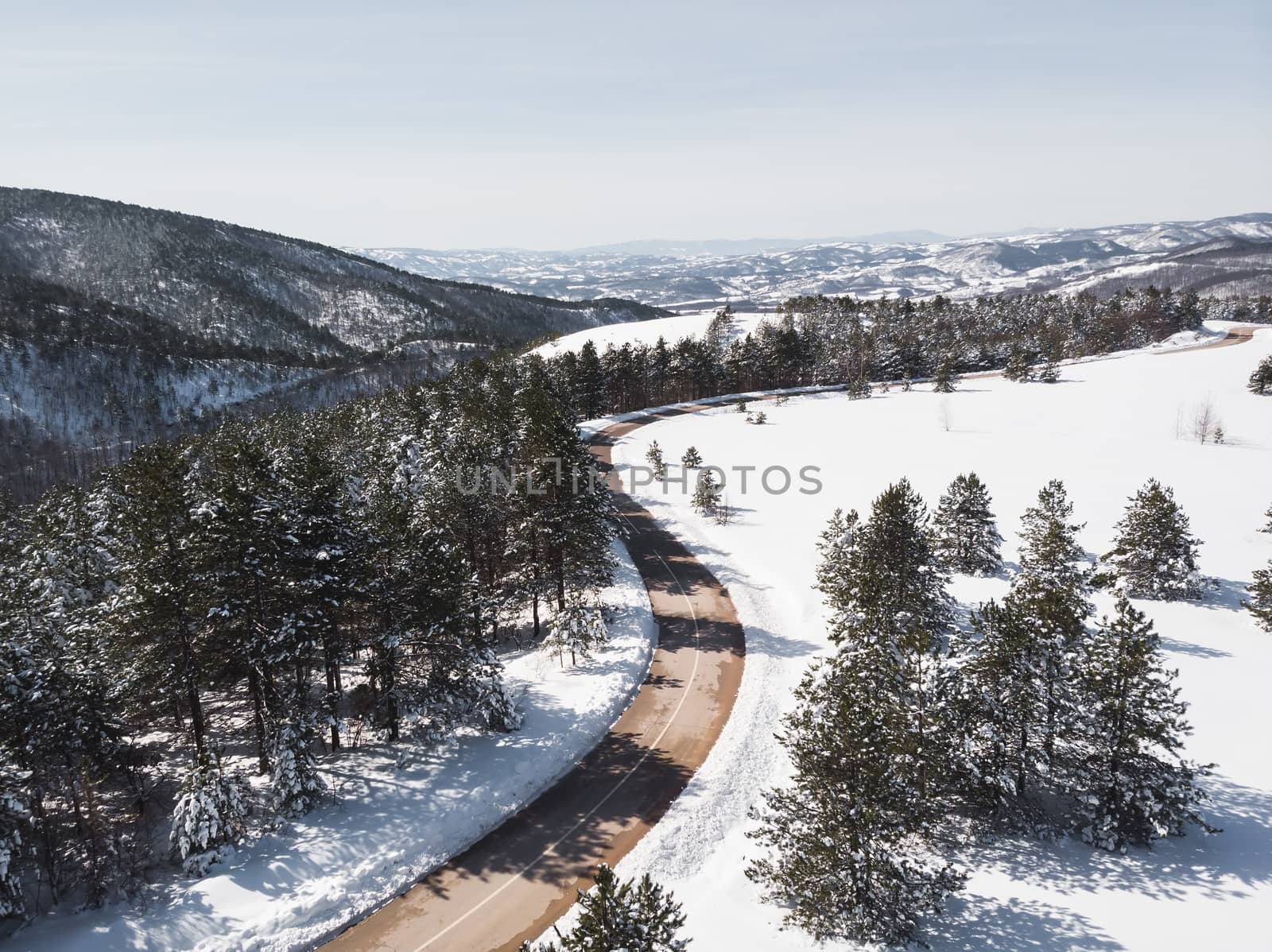 Winter landscape in sunlight. View on the mountain road surrounded by evergreen trees in winter, drone shot