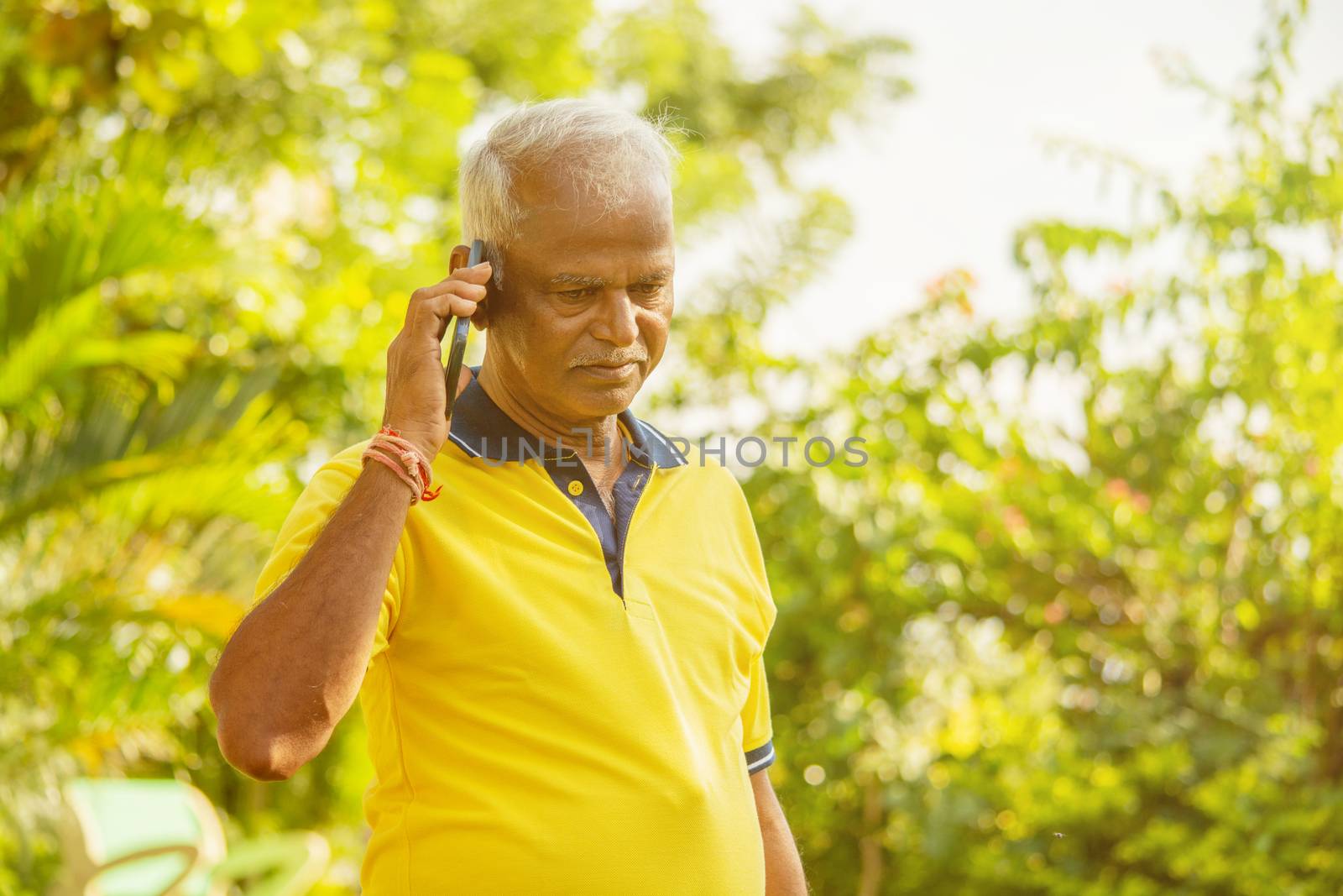 Elder man busy in mobile - Healthy senior Indian on phone outdoor at park, Morning - Happy male parent talking on phone