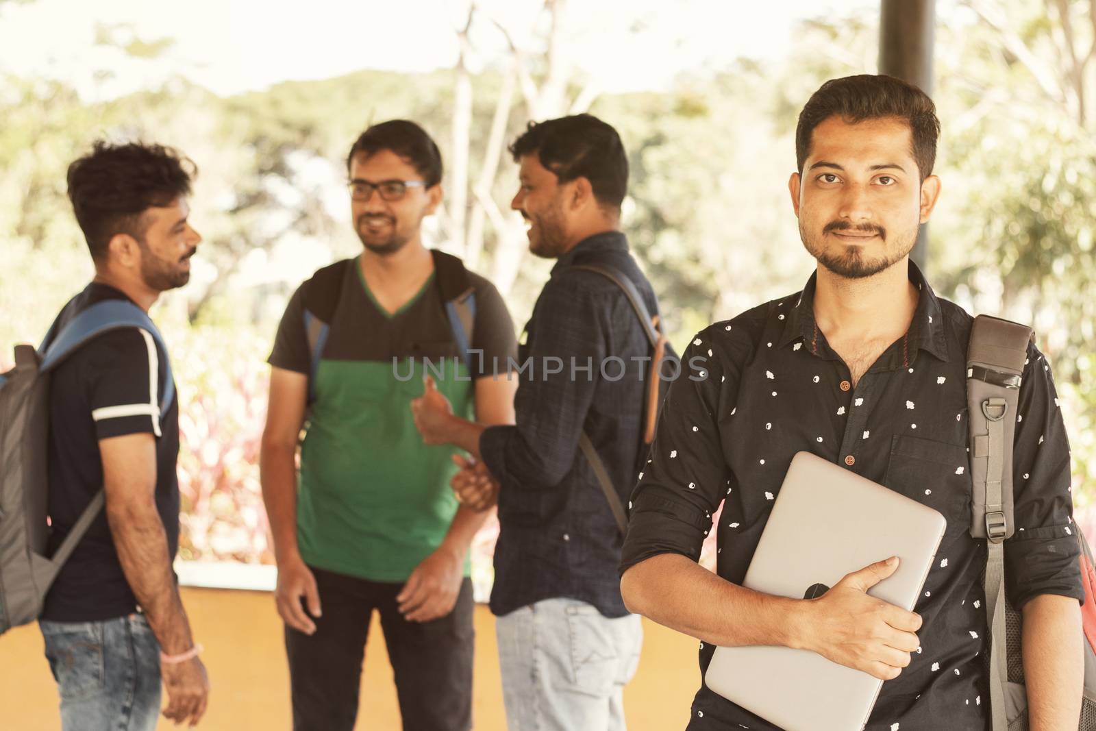 Smiling young adult student with holding laptop in hand while friends staning in background - Concfident graduate scholar smiling at university college