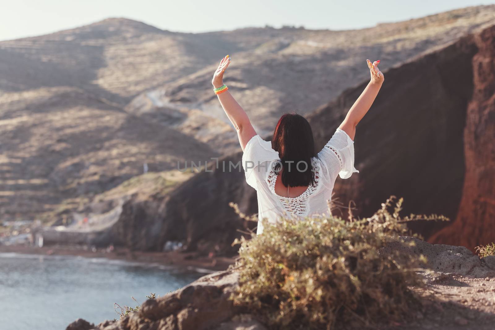 Back view of happy woman on the beach with hands up, sitting on rocks of famous red beach, with volcanic sand  and  rocky shoreline, Santorini, Akrotiri, Greece